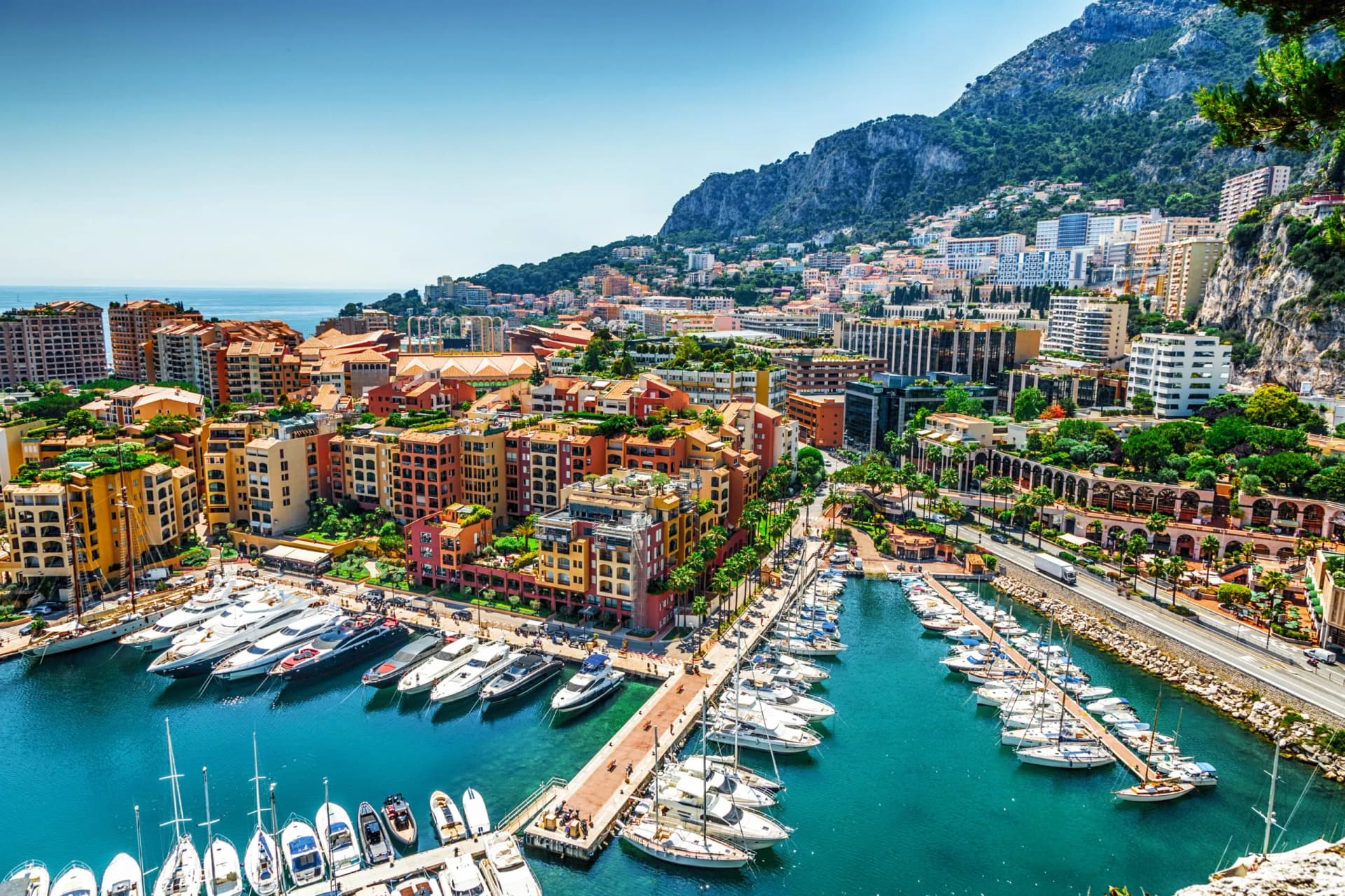 Yachts and sailboats docked in a harbor below colorful buildings and a steep, green mountain.