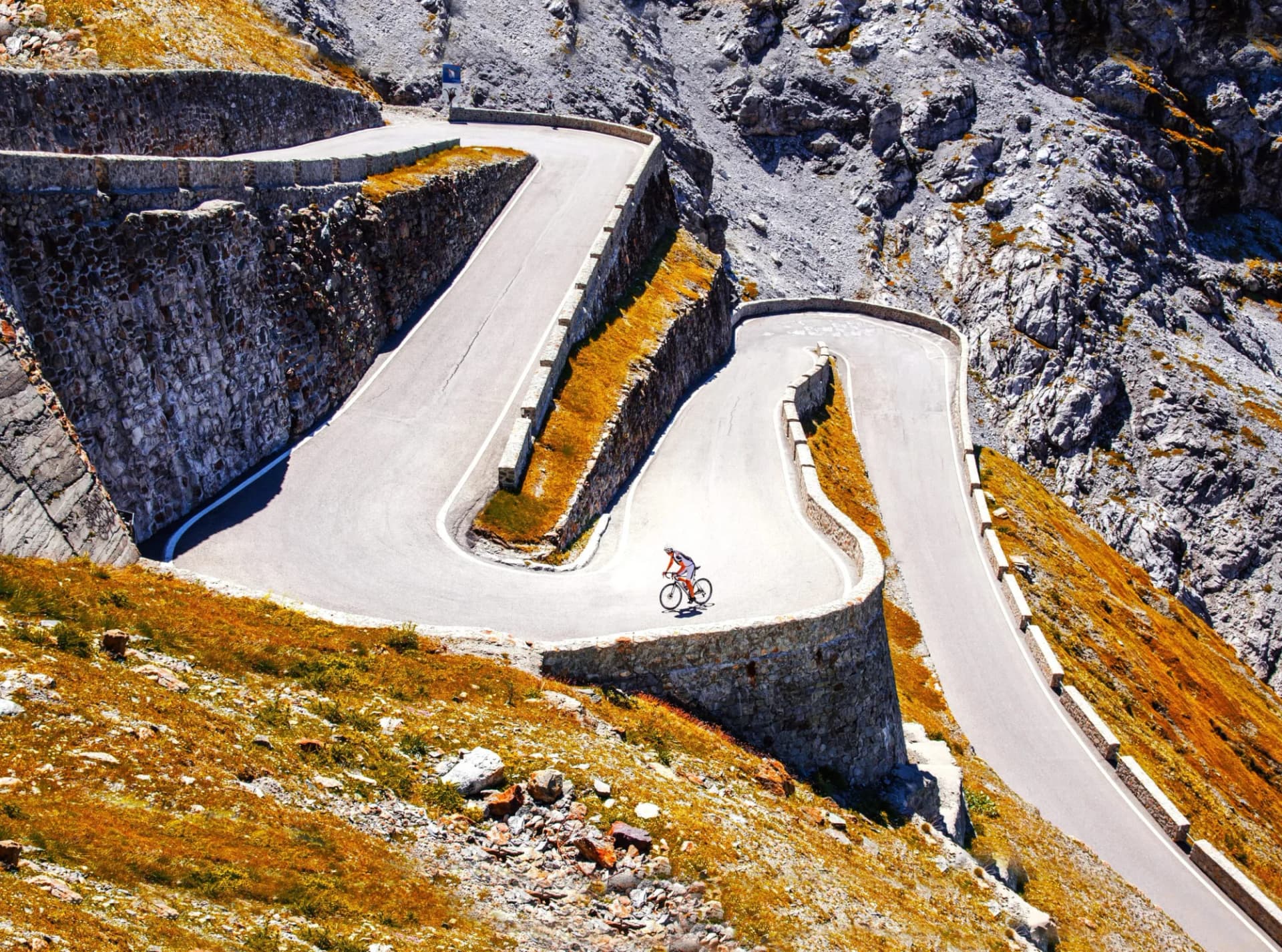 Cyclist ascending steep alpine switchback road with stone retaining walls and dry grass.