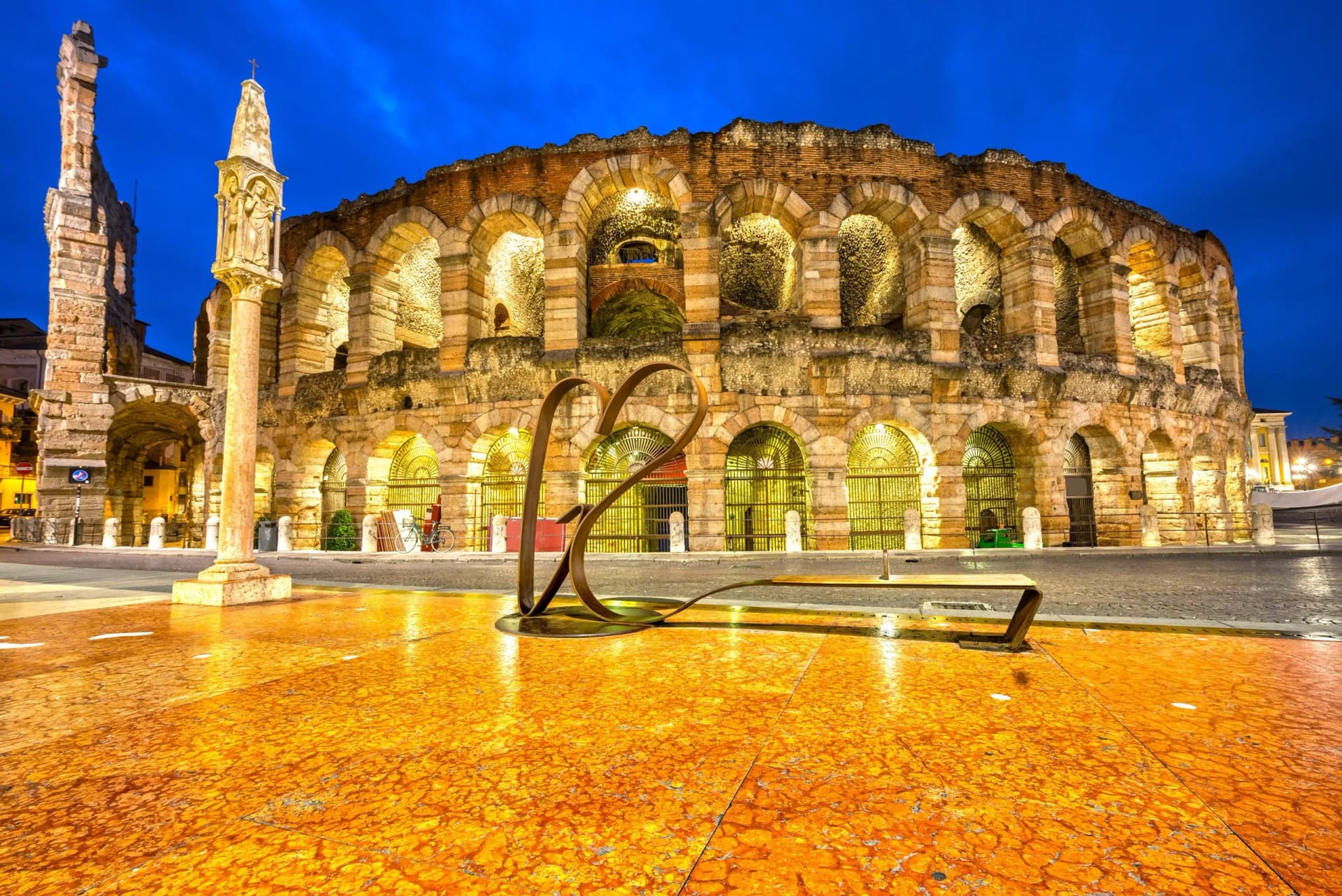 Illuminated Verona Arena at dusk with a heart sculpture and stone column in the foreground.