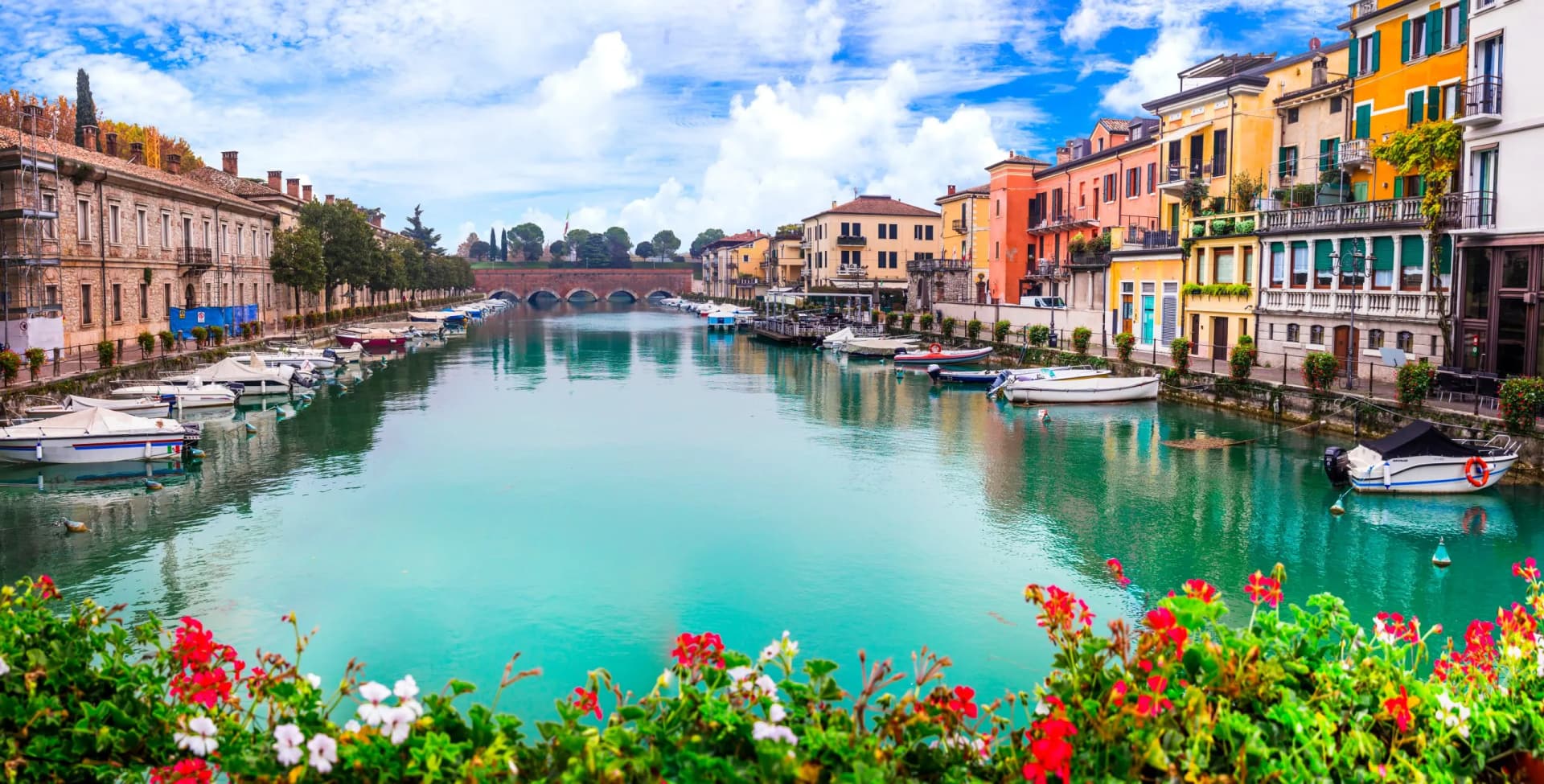 Boats docked on turquoise canal beside colorful European buildings, viewed over red flowers.