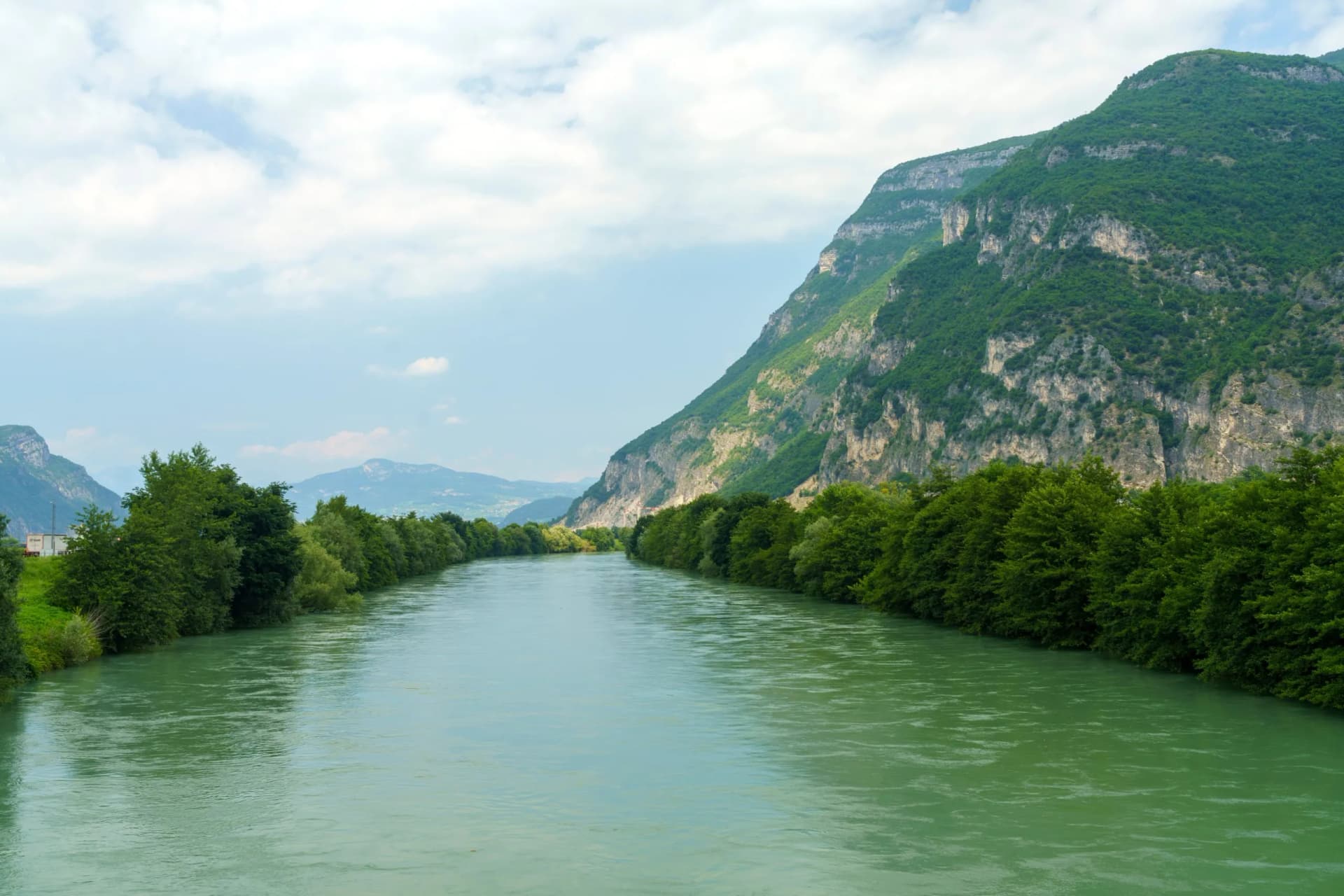 Wide green river flowing between tree-lined banks and steep, rocky mountains under a cloudy sky.
