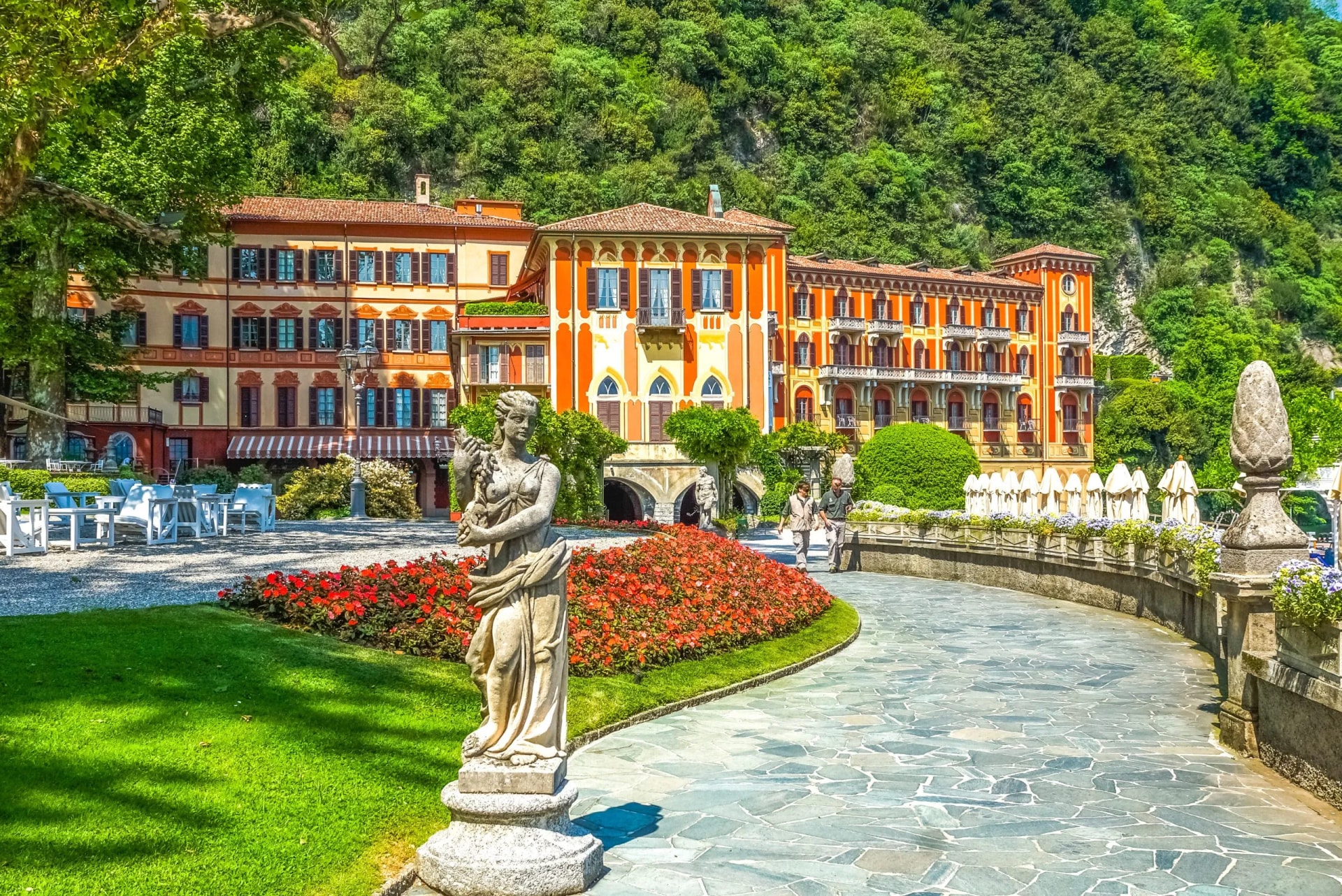 Statue in garden with red flowers, colorful hotel, and lush green mountain backdrop.