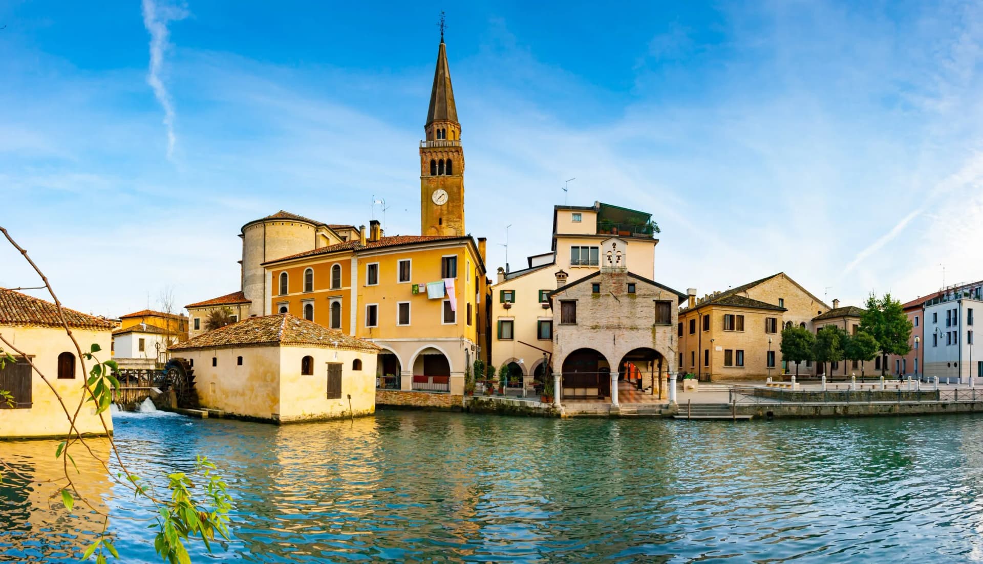 Waterfront buildings with a tall clock tower reflected in the rippling water of Portogruaro.