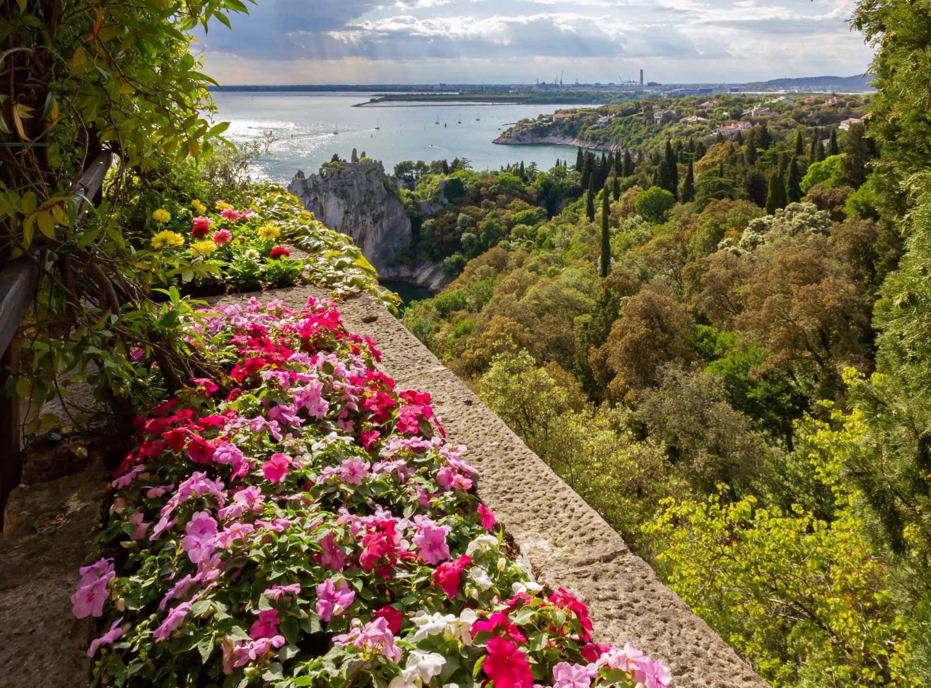 Flower-lined stone balcony overlooking a coastal bay with forested hills and distant industrial cranes.