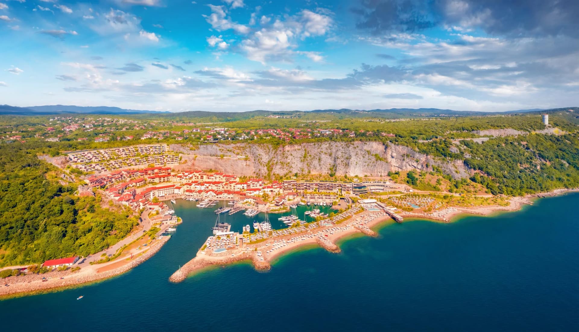 Coastal resort town with red-roofed buildings nestled against a cliff overlooking a marina filled with yachts.
