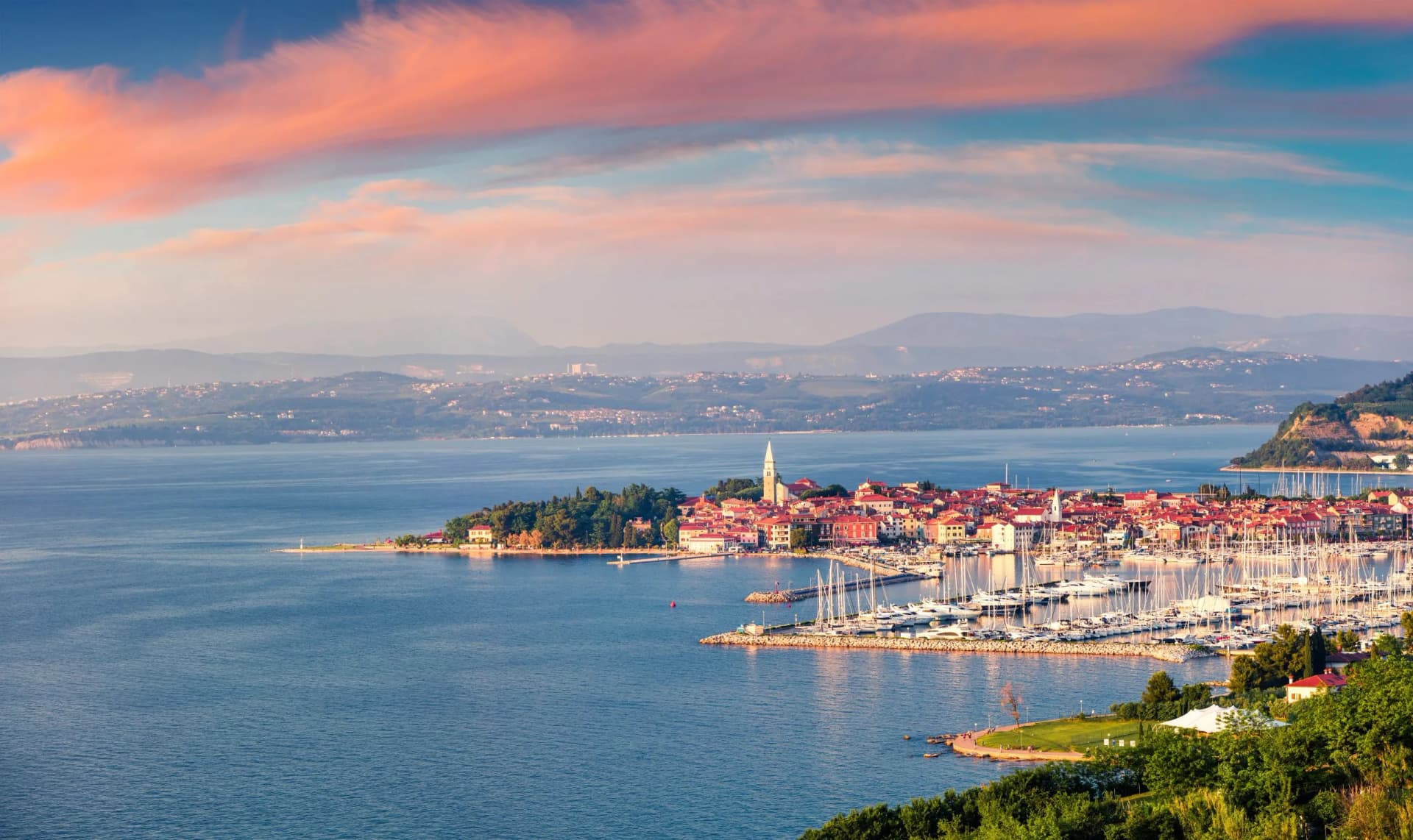 Coastal town with red-roofed buildings and marina full of sailboats at sunset, Izola, Slovenia.
