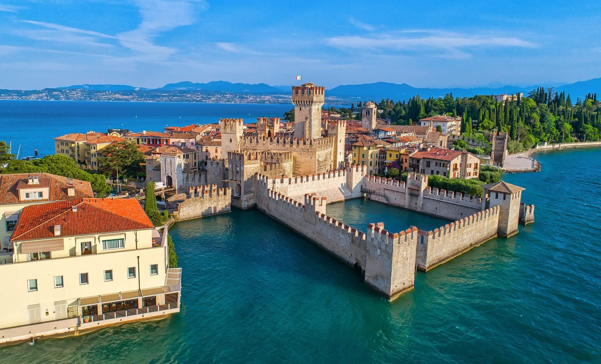 Scaliger Castle in Sirmione surrounded by turquoise water of Lake Garda with mountains in background.