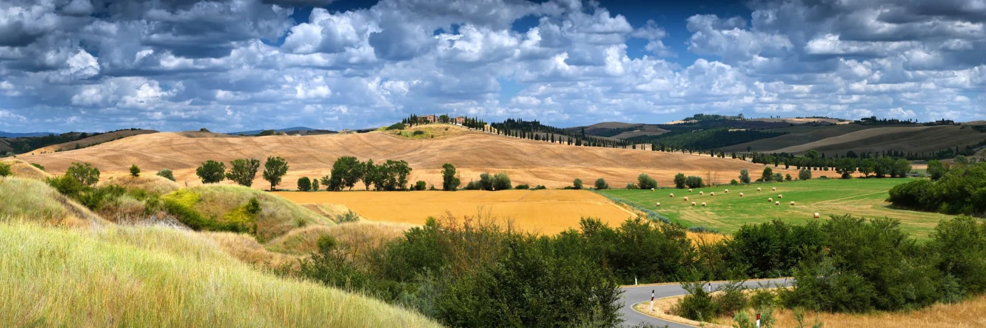 Rolling hills of Tuscany with golden fields, cypress trees, and hay bales under dramatic clouds.
