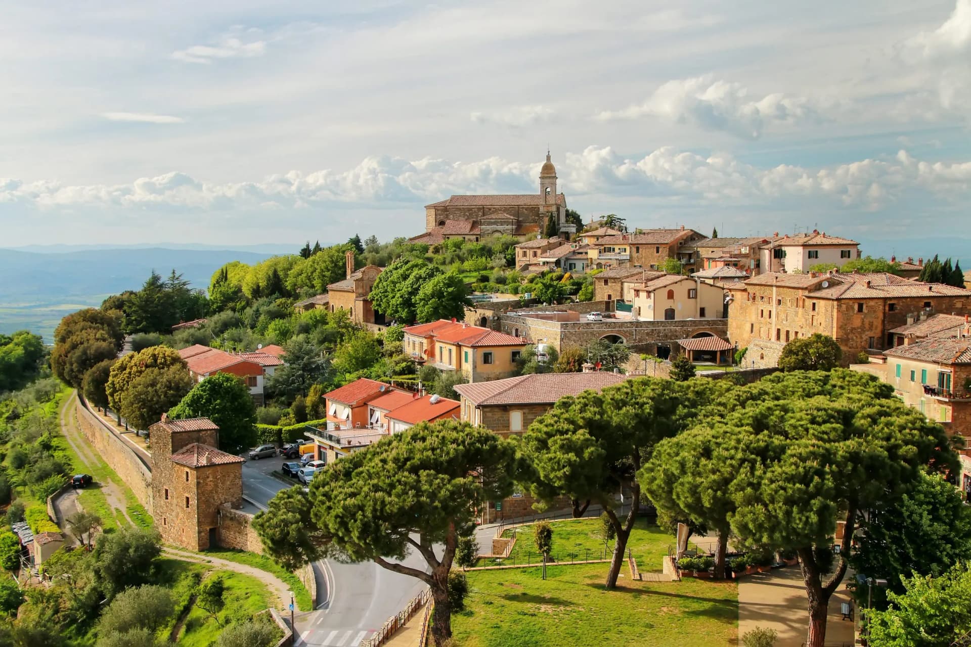 Hilltop town with terracotta roofs, church tower, and green trees under a cloudy sky.