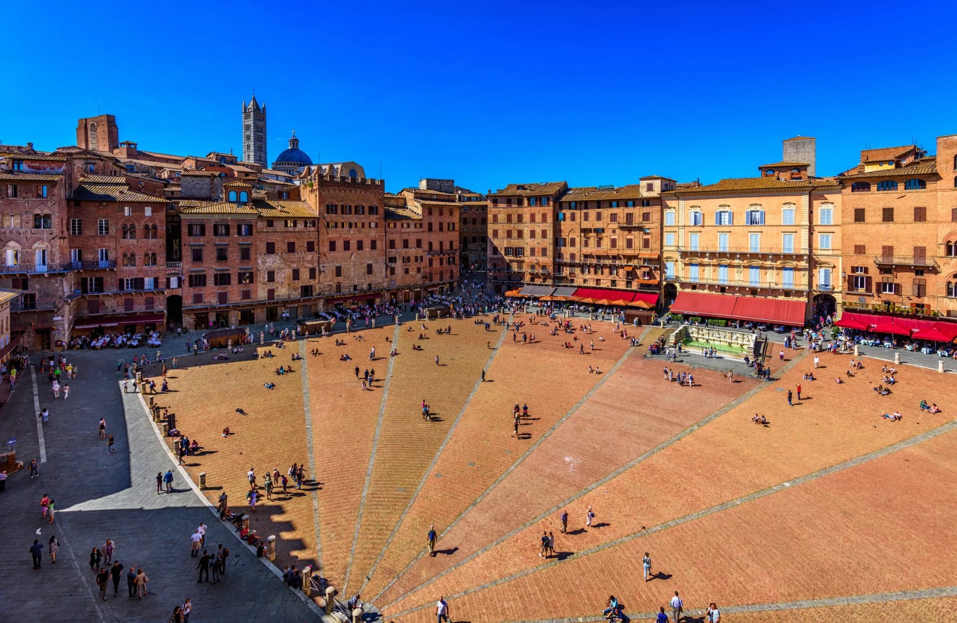 Piazza del Campo with people relaxing on the brick pavement and historic buildings under a clear blue sky in Siena.