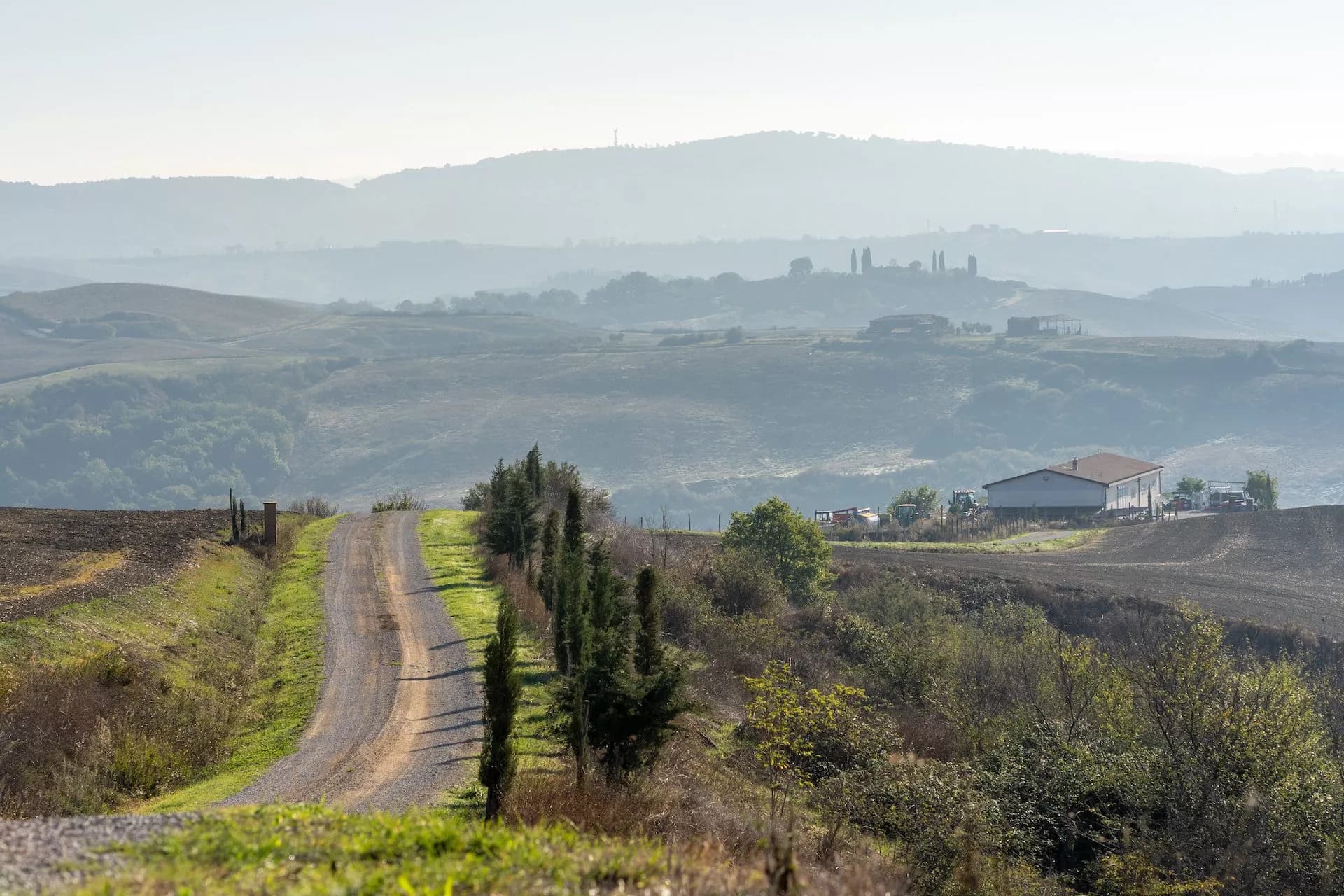 Gravel road through rolling hills with cypress trees, farm buildings, and hazy mountains in Tuscany.