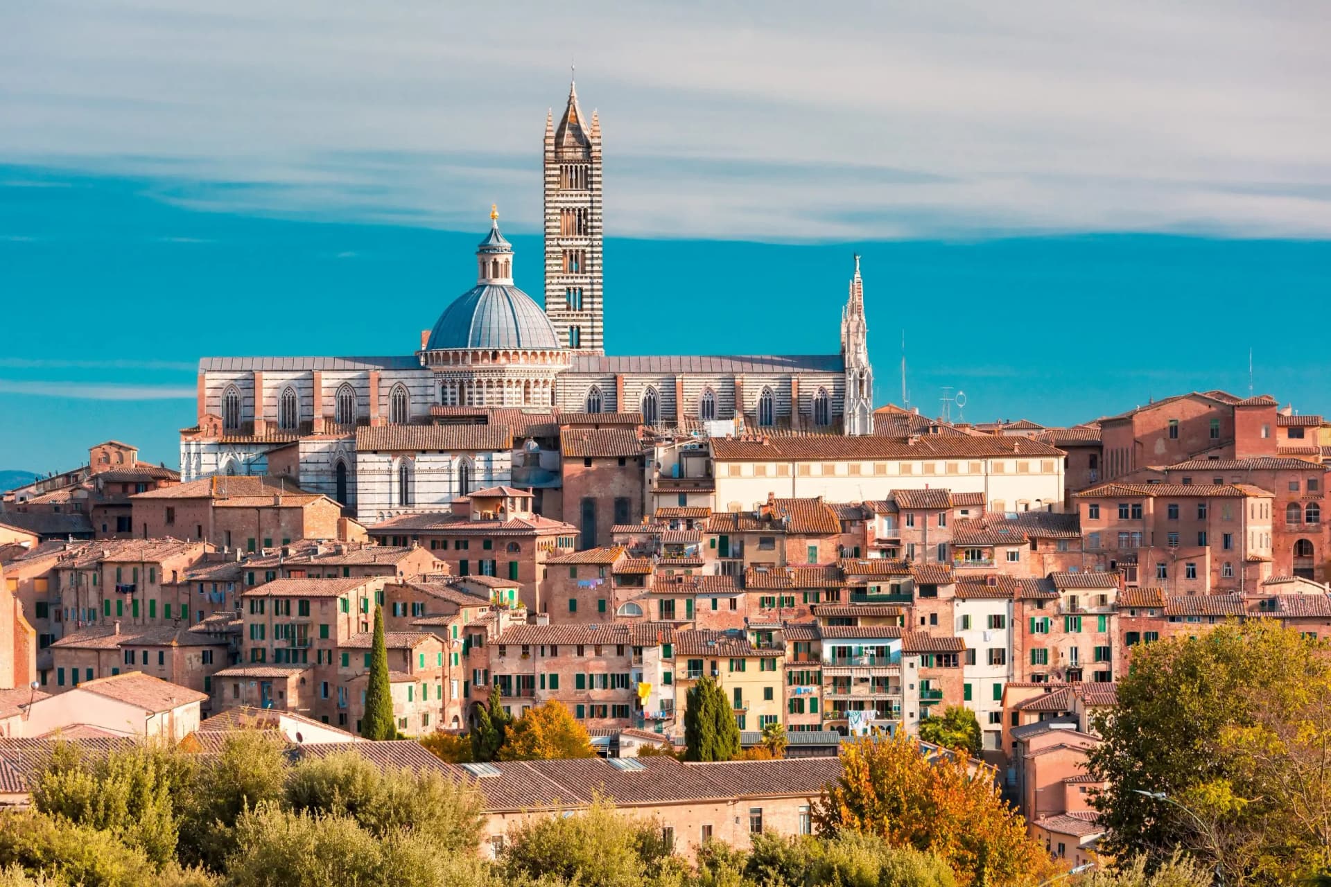 Siena skyline with Duomo dome and striped bell tower above terracotta rooftops under bright blue sky.