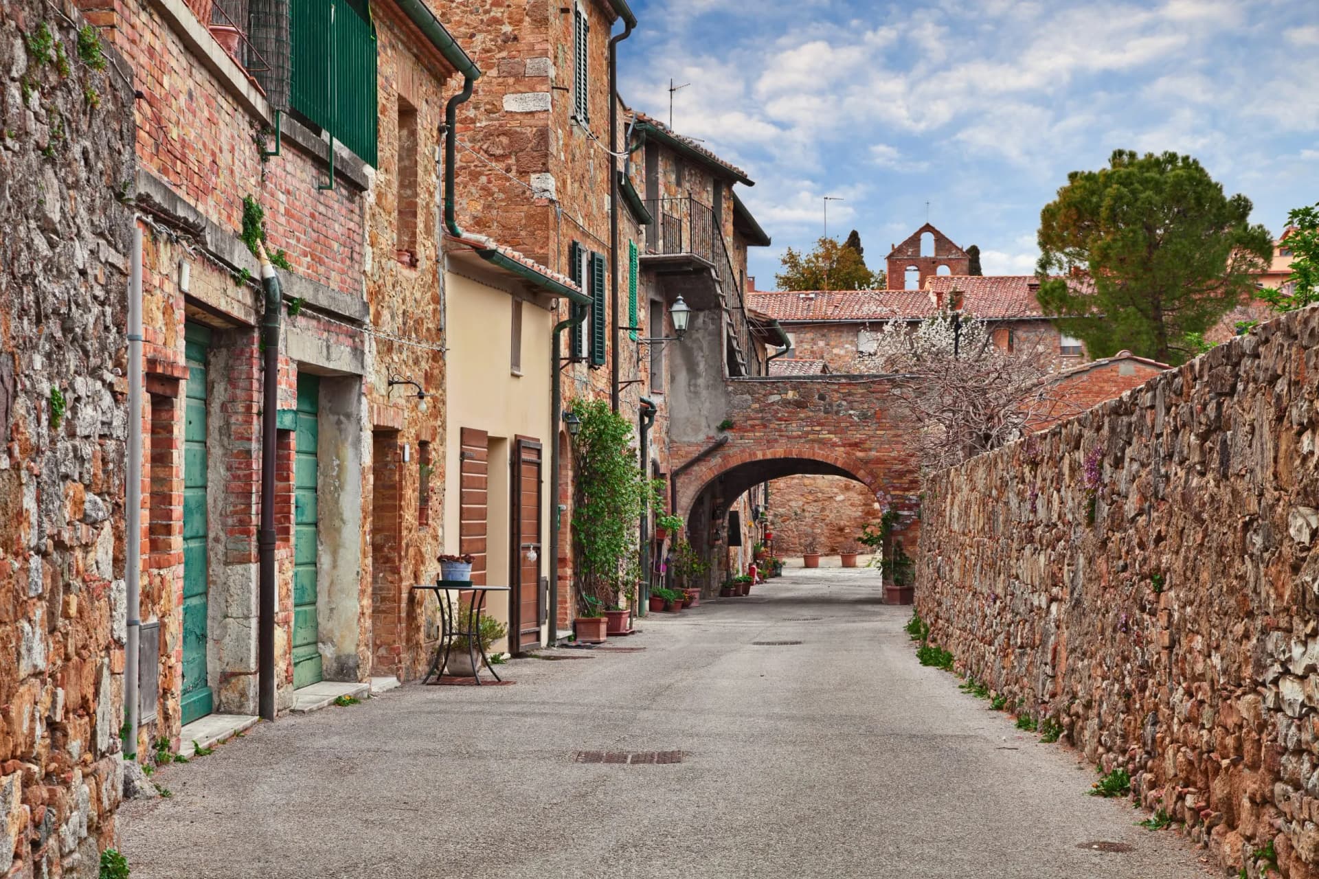Narrow street in Val d'Orcia with stone buildings, an archway, and blooming trees under a cloudy sky.