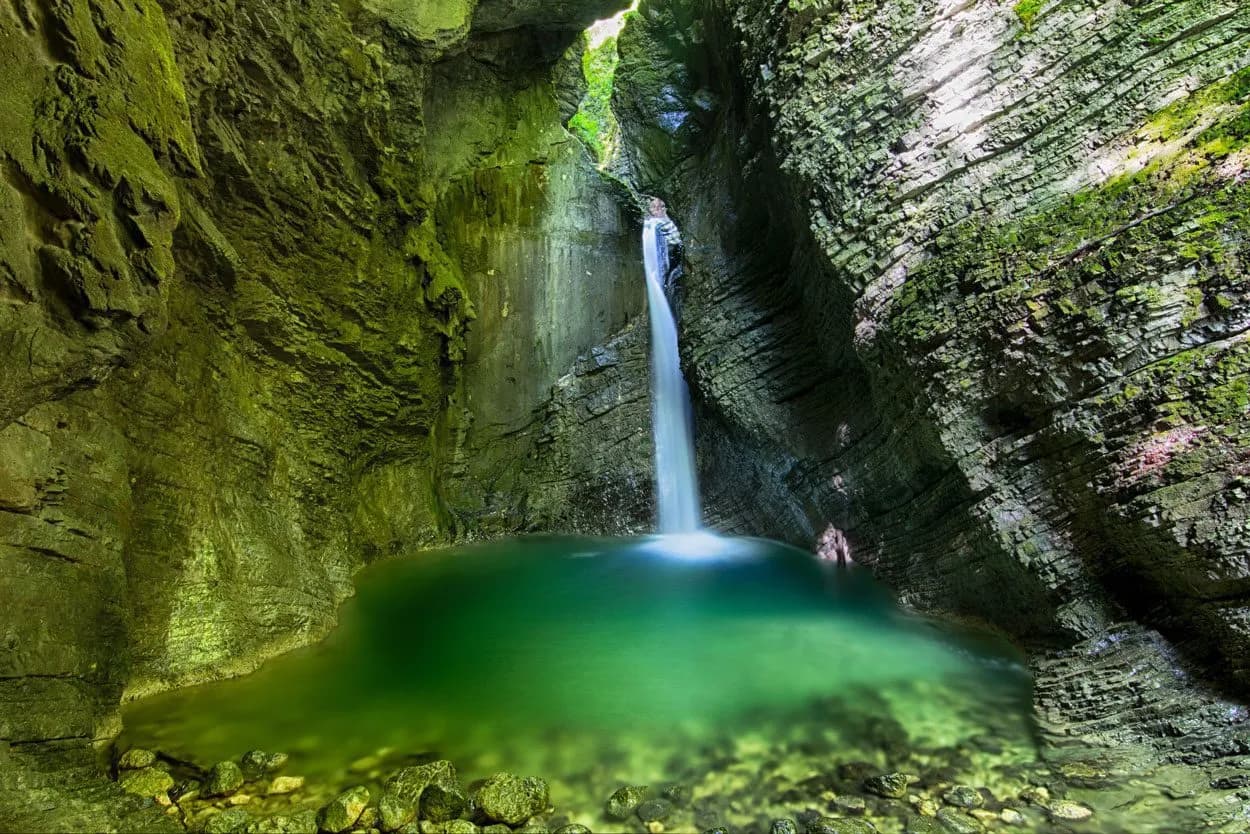 Kozjak waterfall plunging into a deep green pool inside a moss-covered rock canyon.