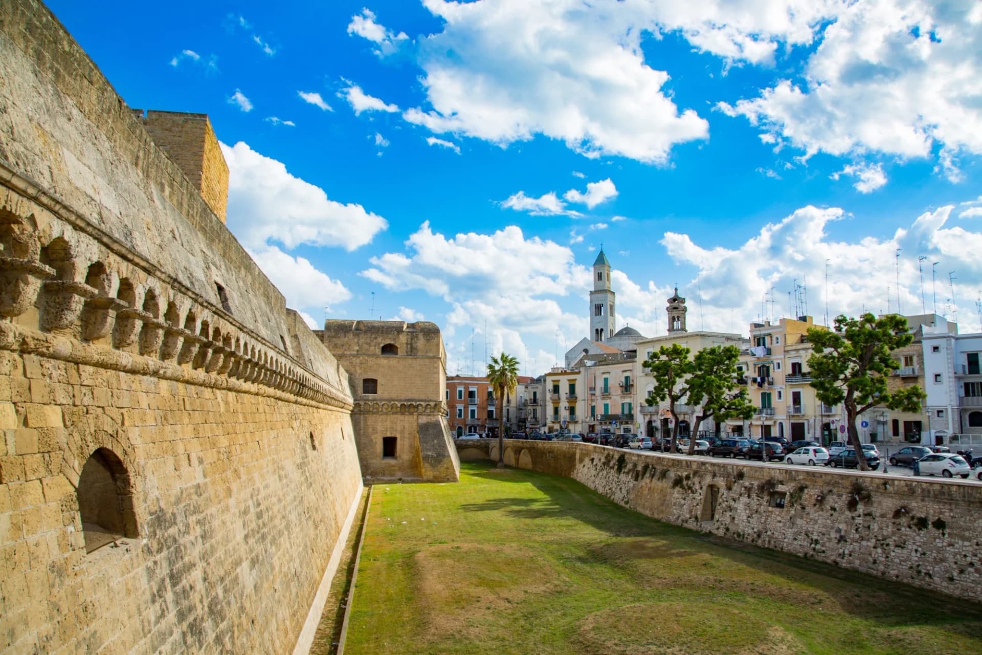 Stone fortress wall next to a dry moat with historic buildings and a church tower in Bari.