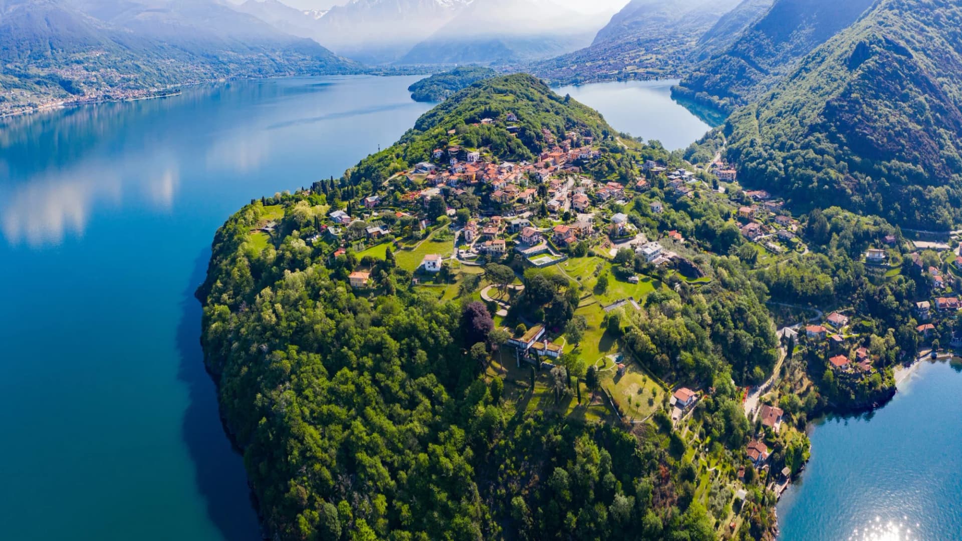 Aerial view of Colico Olgiasca peninsula with village houses surrounded by lush green hills and Lake Como.
