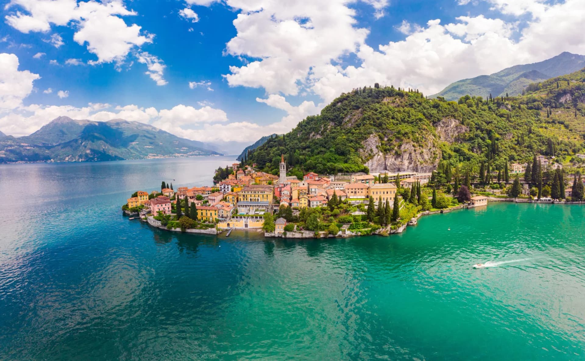 Colorful village on peninsula jutting into turquoise lake surrounded by green mountains under blue sky.
