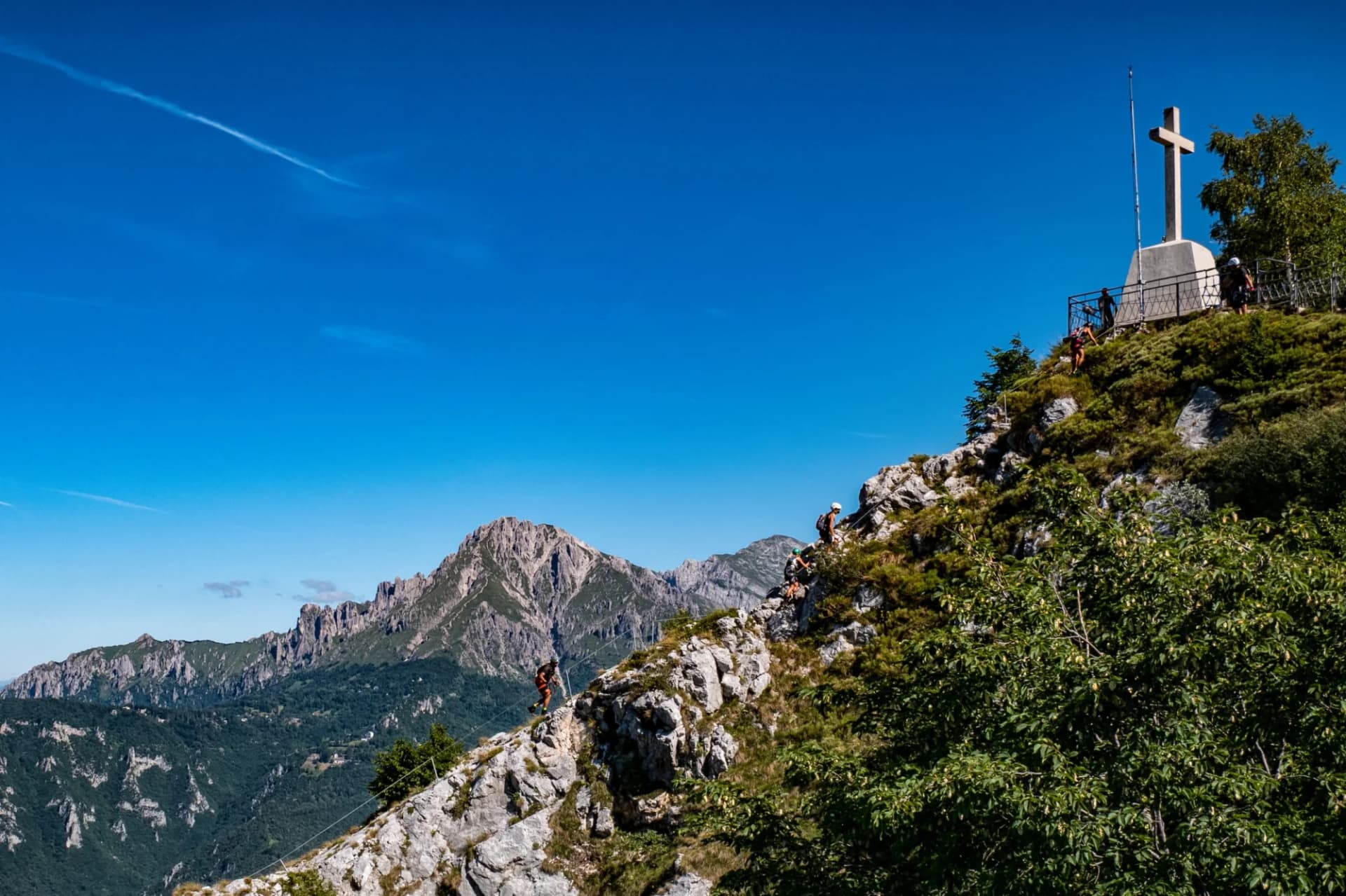Hikers ascending rocky terrain toward a summit cross with Grigna mountain in the background.
