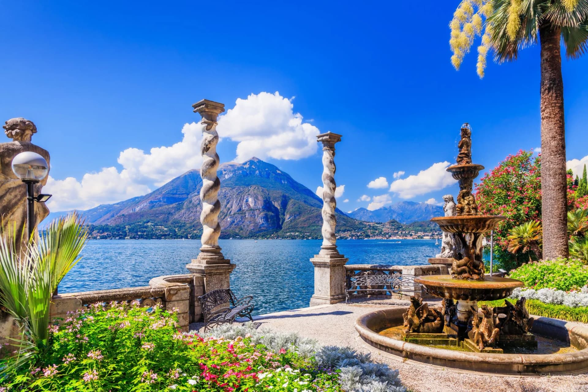 Terrace with ornate fountain, spiral columns, and palm trees overlooking a mountain lake.