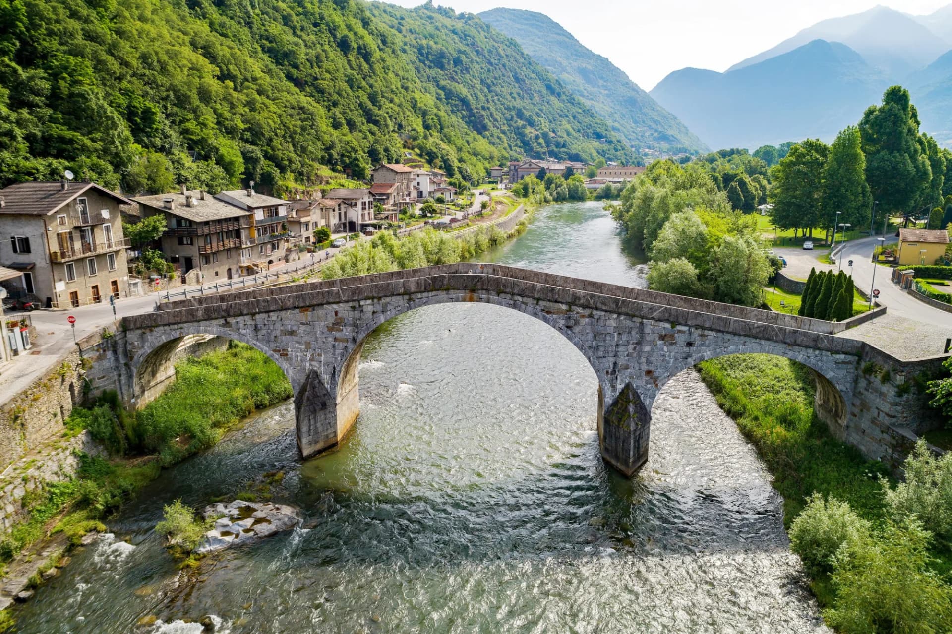 Stone arch bridge over a river in an alpine village setting with lush green mountains.