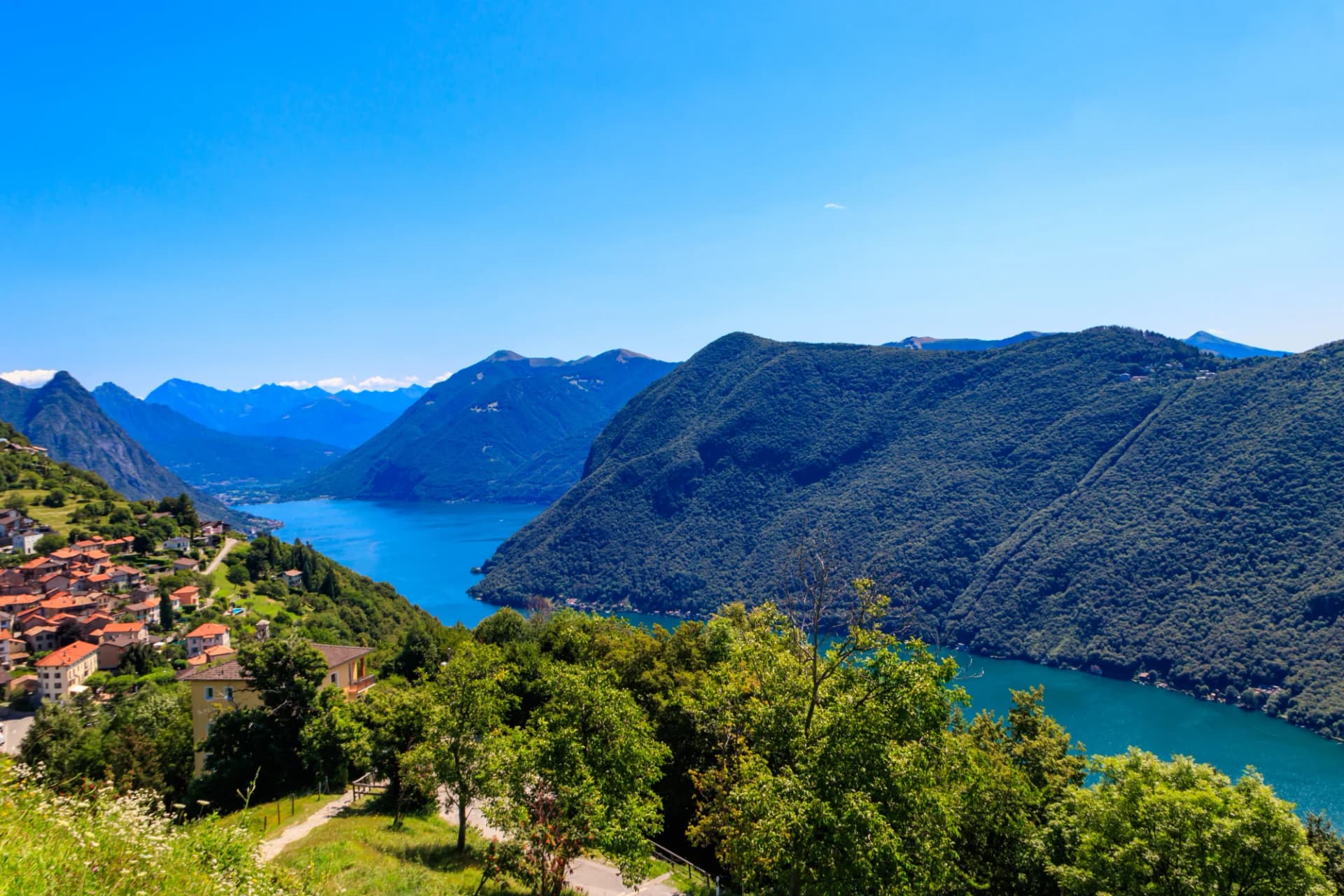 Village on hillside overlooking Lake Lugano with steep, forested mountains under blue sky.