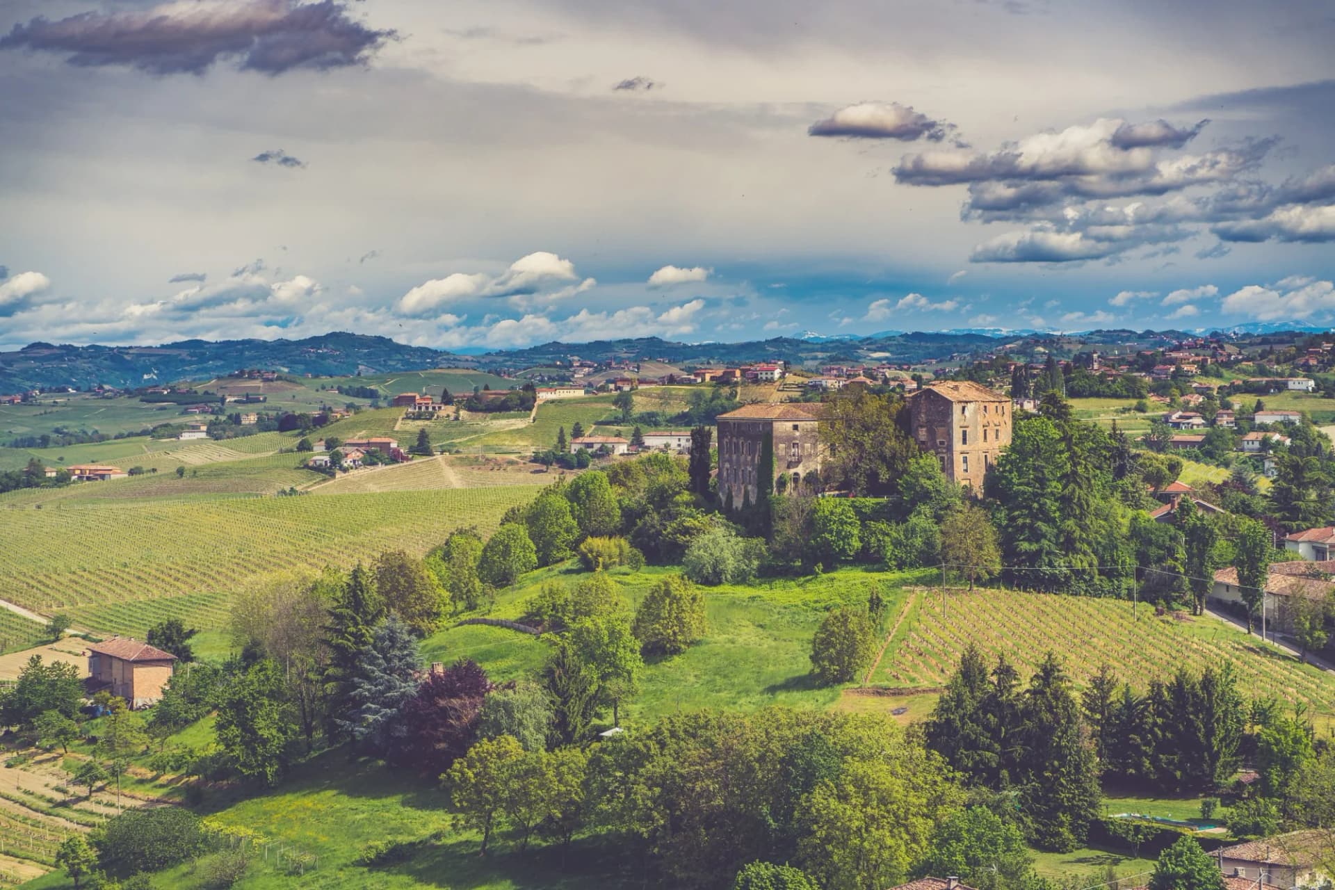 Vineyard landscape with rolling hills, historic stone building, and cloudy sky in Langhe-Monferrato, Piedmont, Italy.