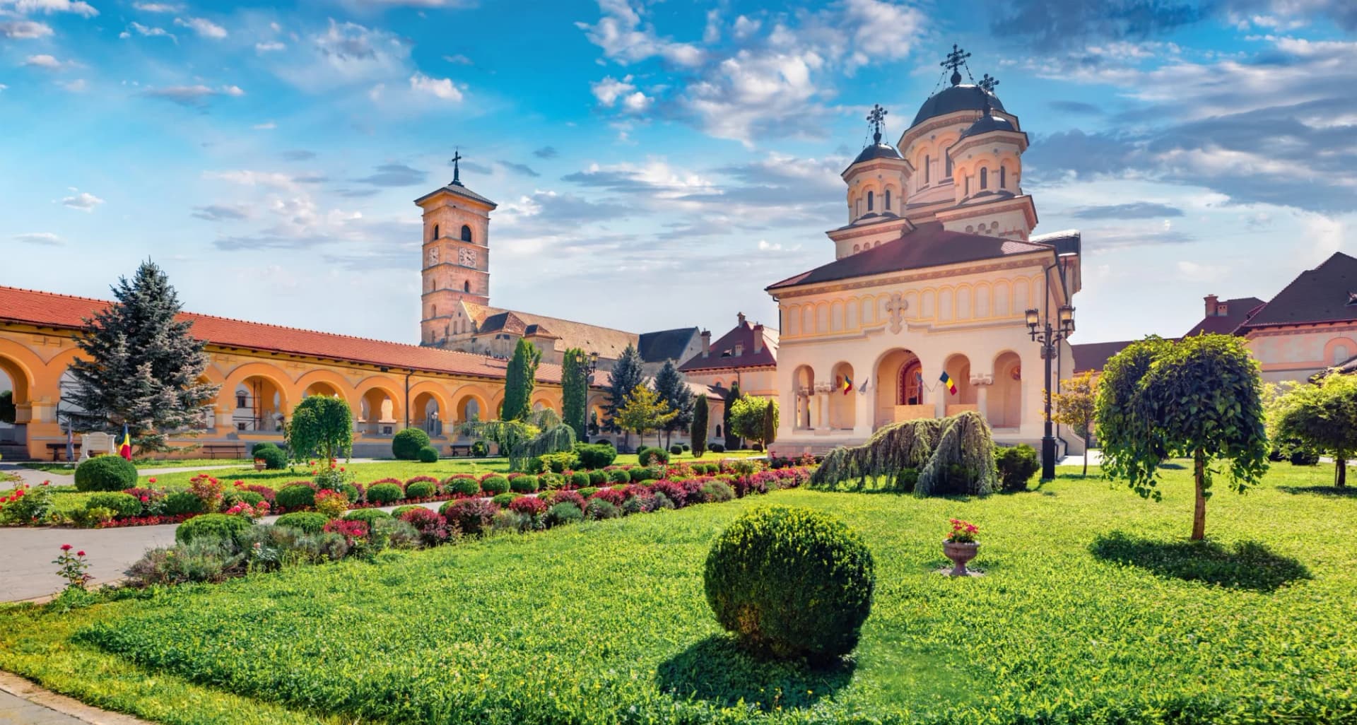 Alba Carolina Fortress courtyard with Orthodox cathedral, clock tower, and manicured gardens under blue sky.