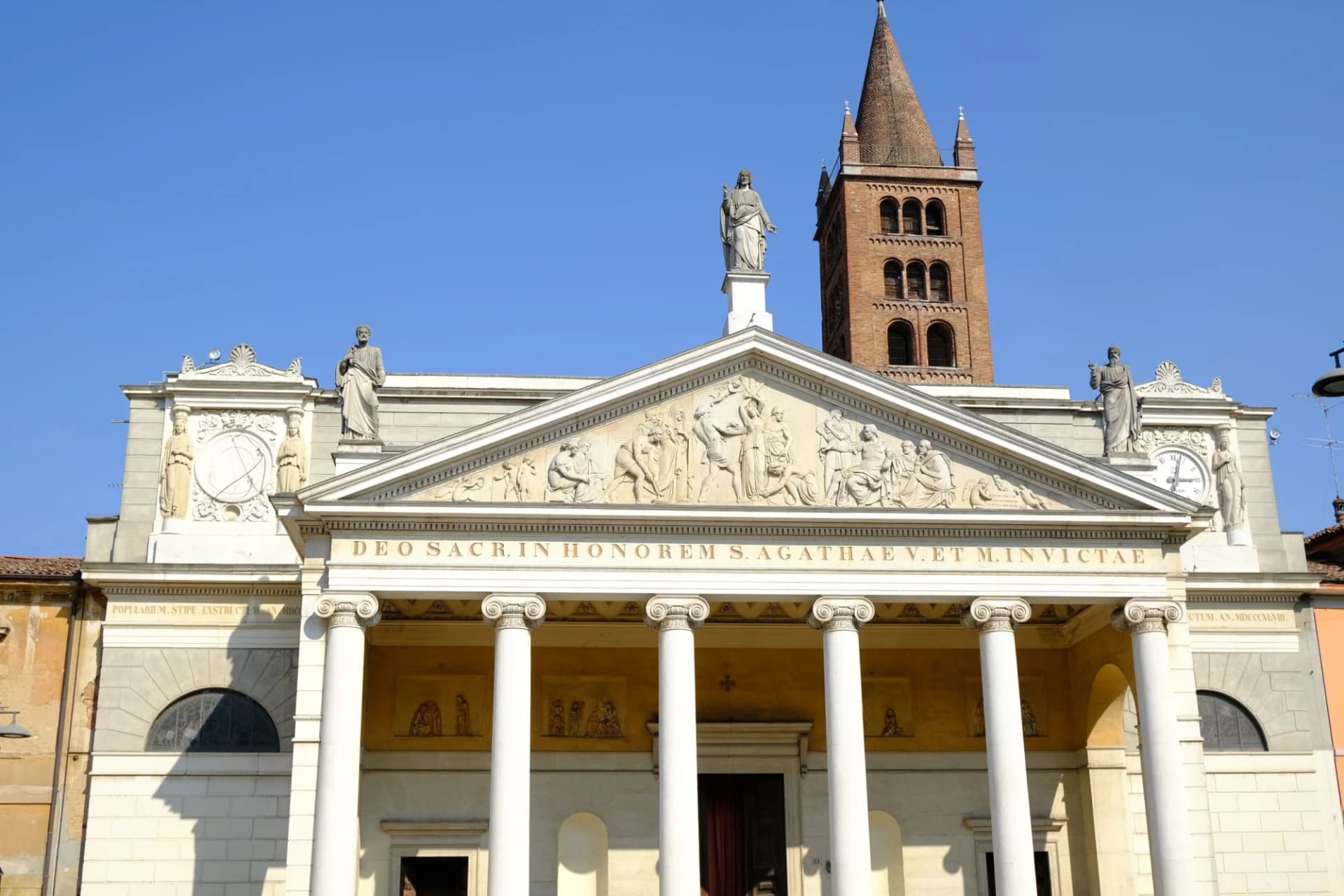 Neoclassical church facade with columns, pediment relief, statues, and a tall brick bell tower under blue sky.