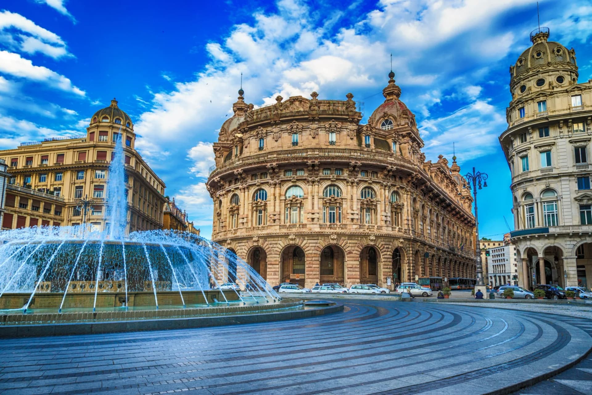 Fountain spraying water in Piazza De Ferrari, Genoa, with ornate historic buildings under a blue sky.