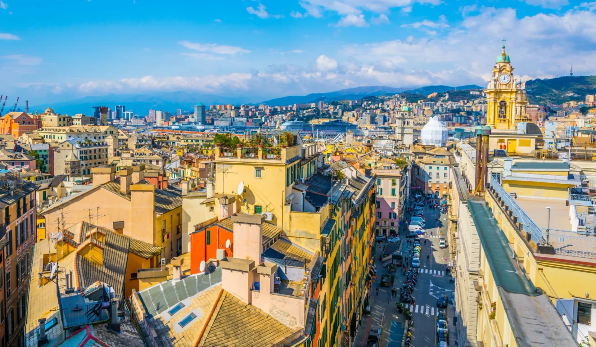 Aerial view of Genoa rooftops, clock tower, and city skyline against blue sky.