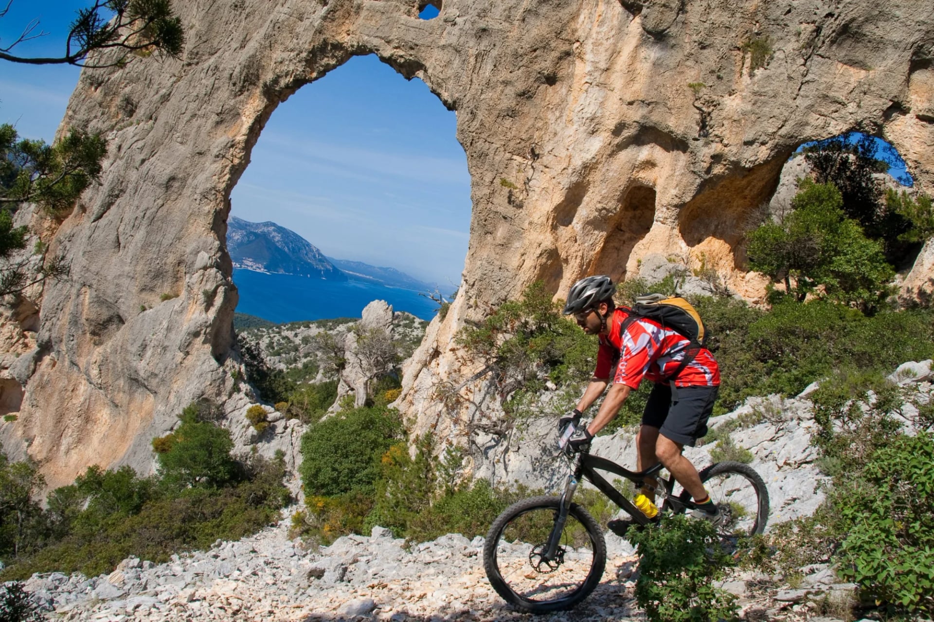 Mountain biker riding near a large natural rock arch overlooking the sea in Sardinia.