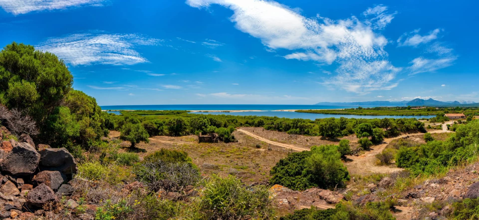 Coastal landscape with scrubland, lagoon, blue sea, and distant mountains under a bright blue sky.