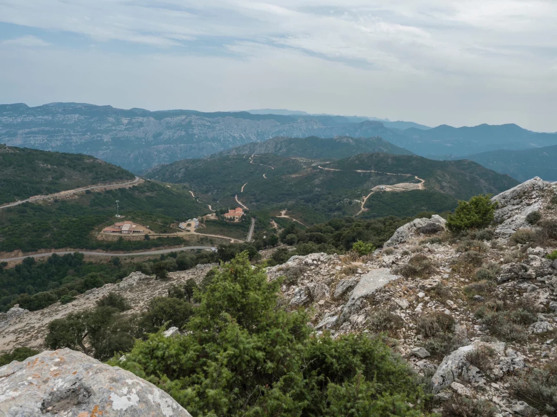 Mountain landscape with winding roads, green hills, and rocky foreground under a cloudy sky