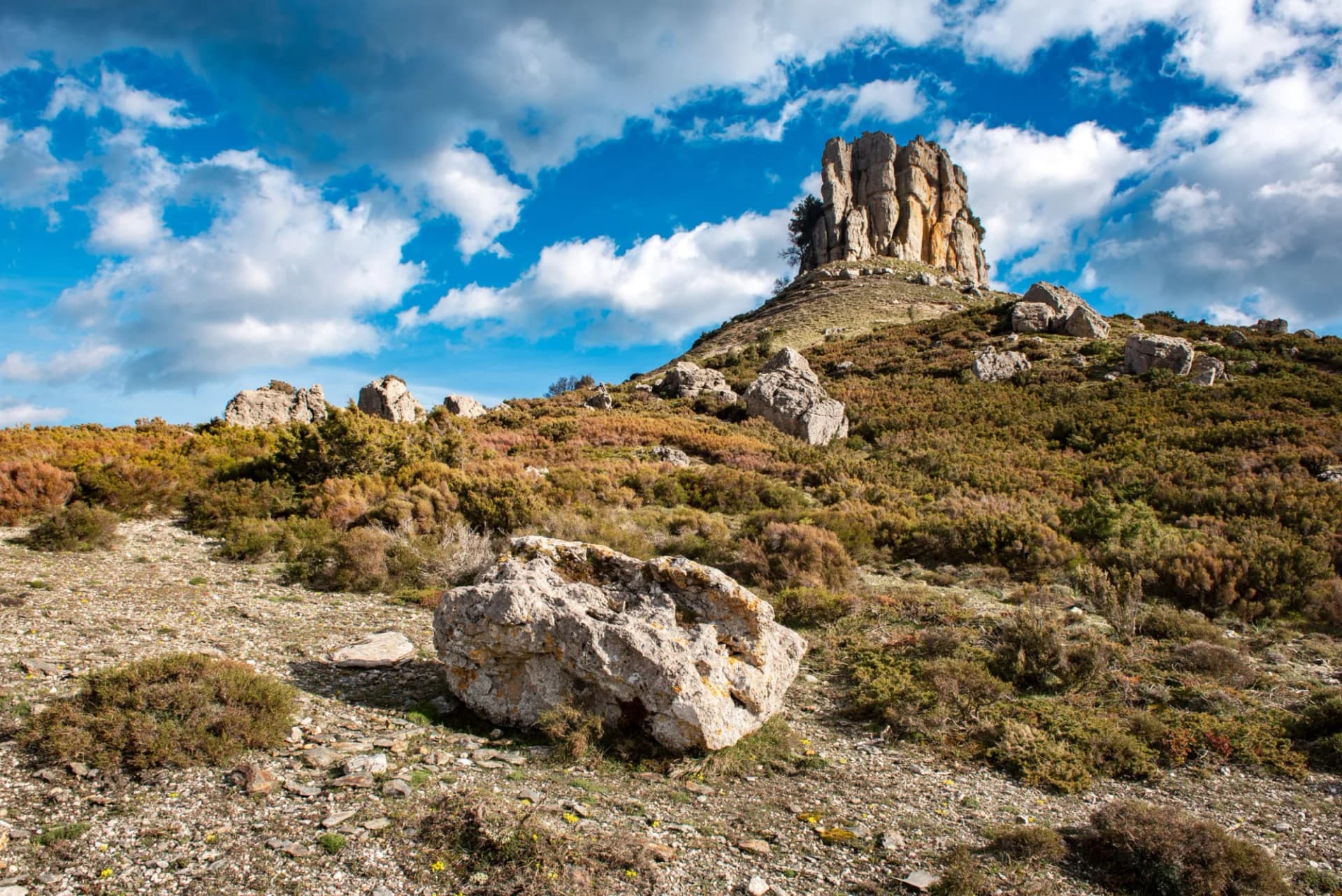 Rocky hillside with scrub brush, large boulders, and a prominent rock formation under a blue, cloudy sky.