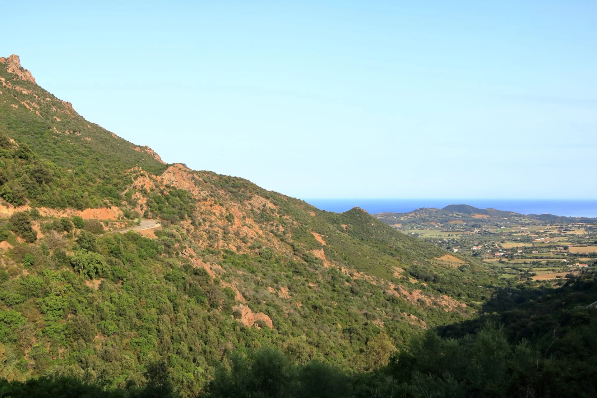 Mountain slope covered in green scrub overlooking a coastal valley and the blue sea.