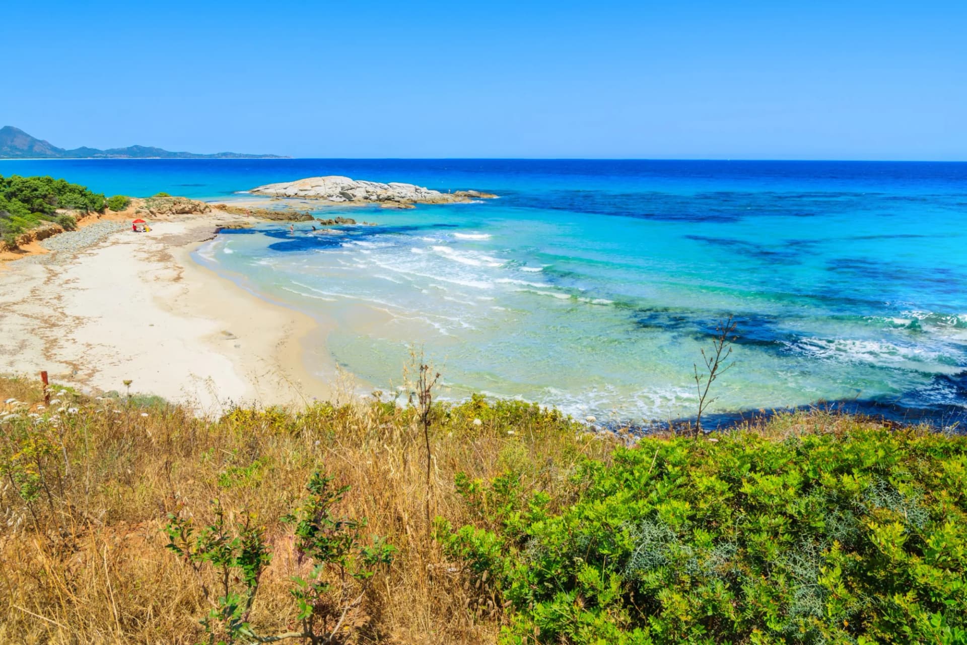 Secluded sandy beach with turquoise water, rocks, and scrubland overlooking the sea.