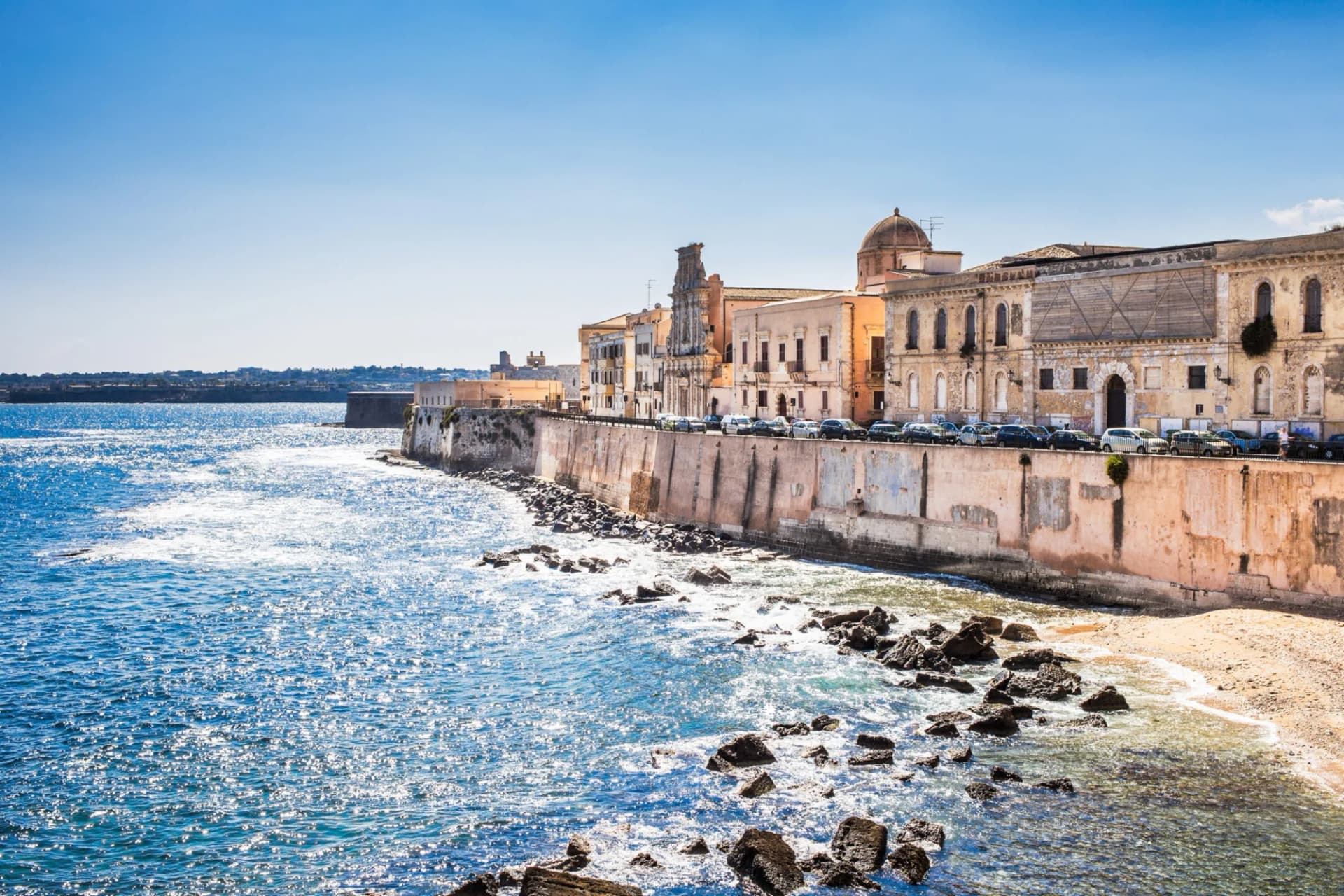 Waterfront historic buildings along the sea wall in Siracusa under a clear blue sky.