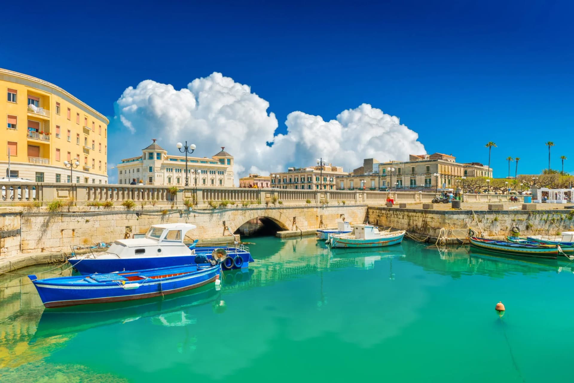 Boats moored in turquoise water beneath a stone bridge in Ortigia, Syracuse, Italy.