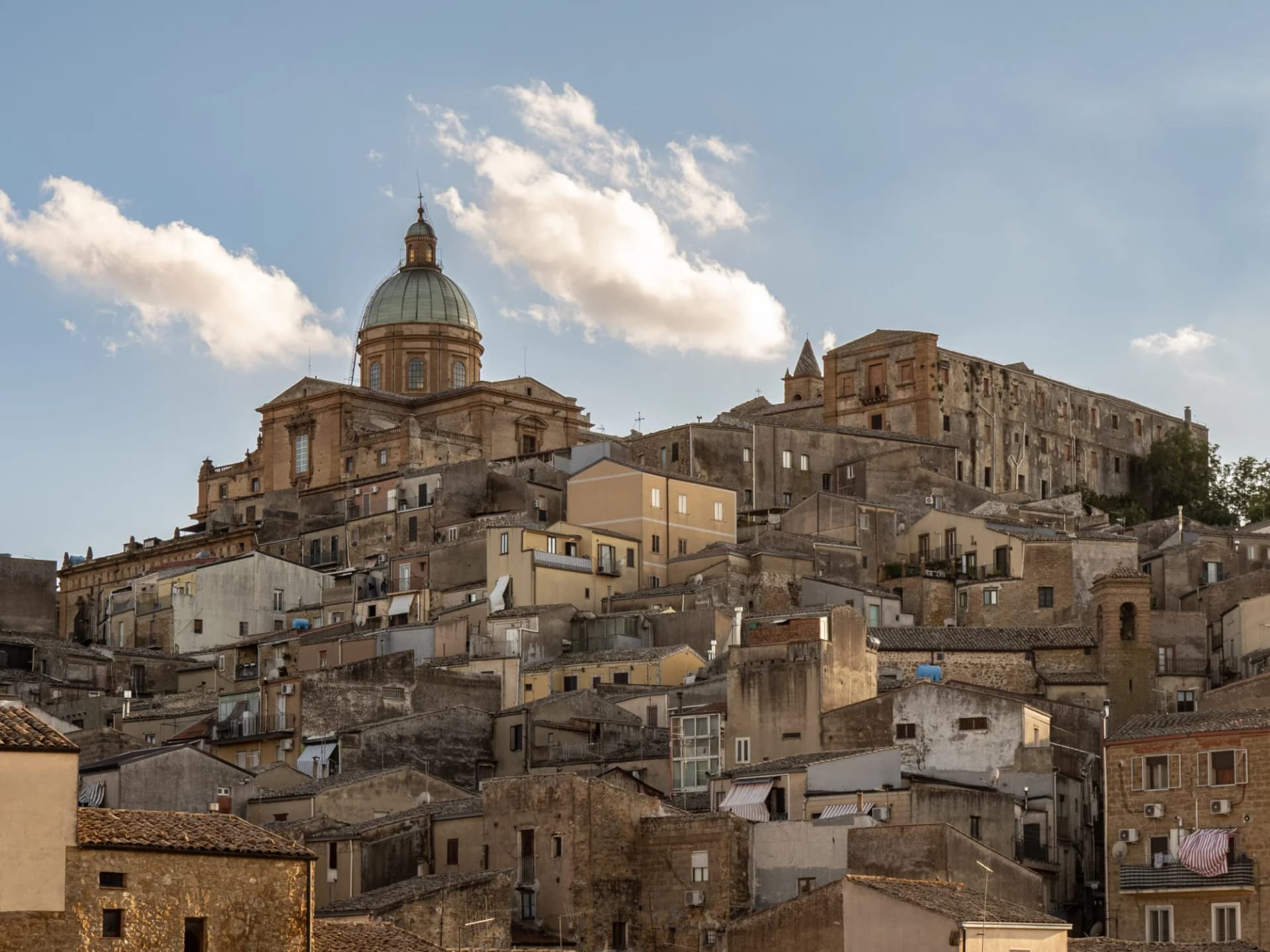 Hillside town with densely packed buildings and a large dome church under a partly cloudy sky, Piazza Armerina.