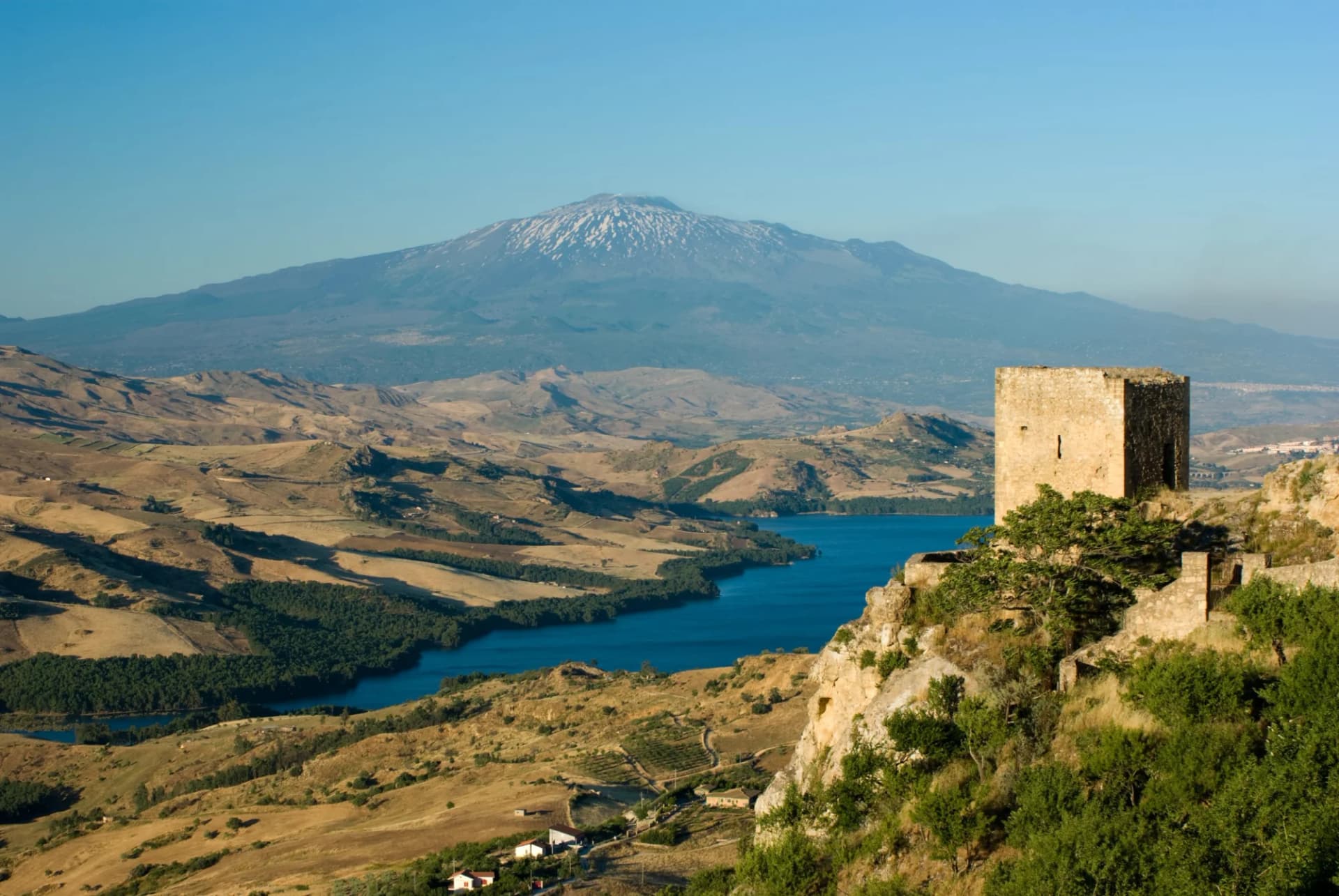 Stone tower ruins overlooking a lake with Mount Etna in the background, Sicily.