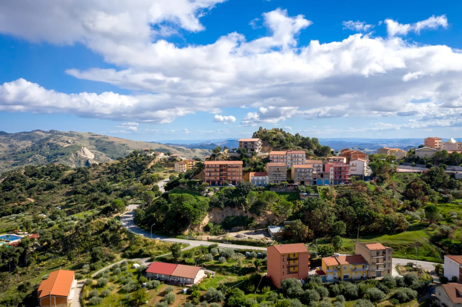 Aerial view of Gagliano Castelferrato, a town on a green hillside under a blue, cloudy sky in Italy.