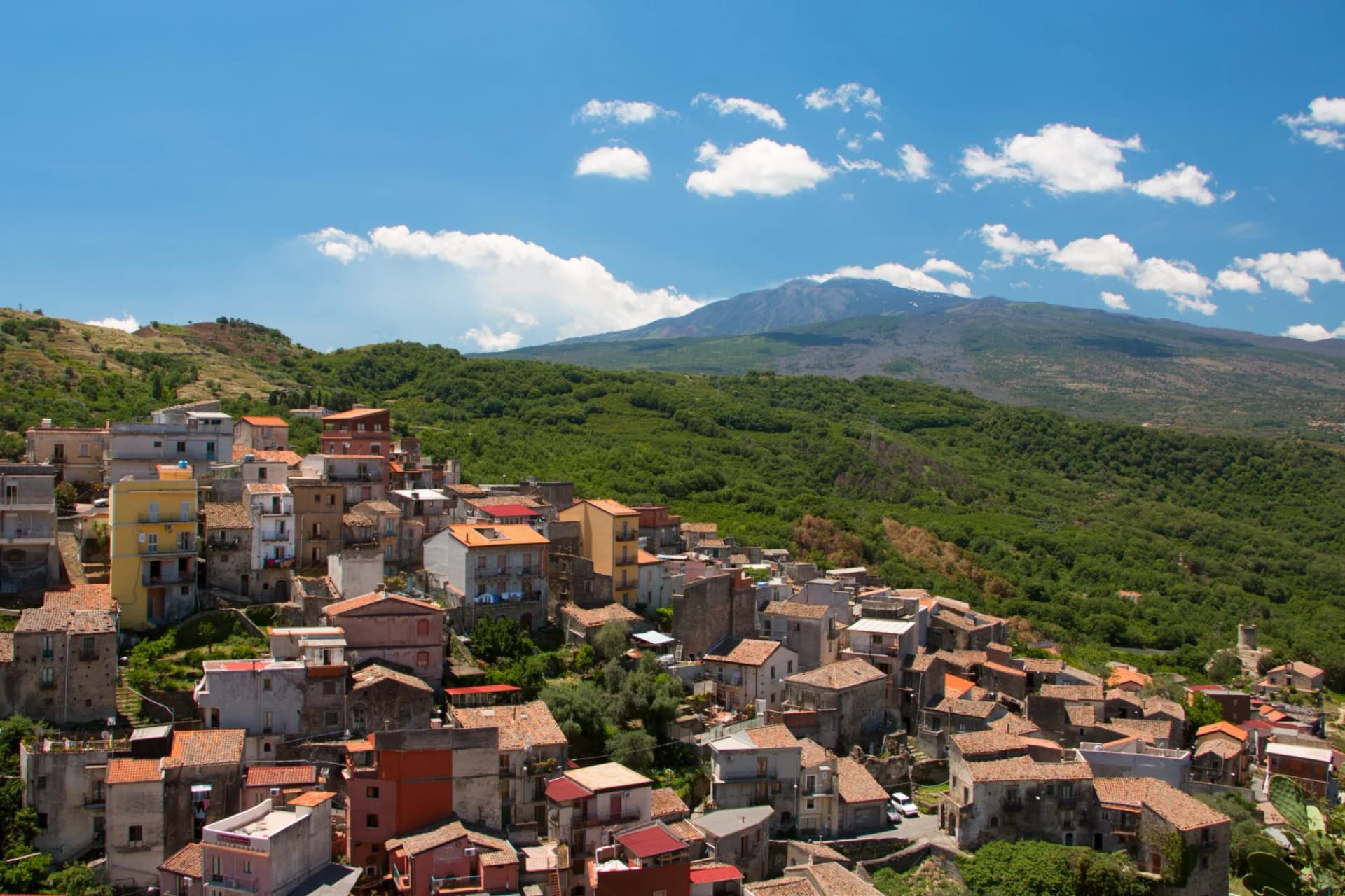 Hillside town with tiled roofs below a large mountain under a blue sky in Sicily.