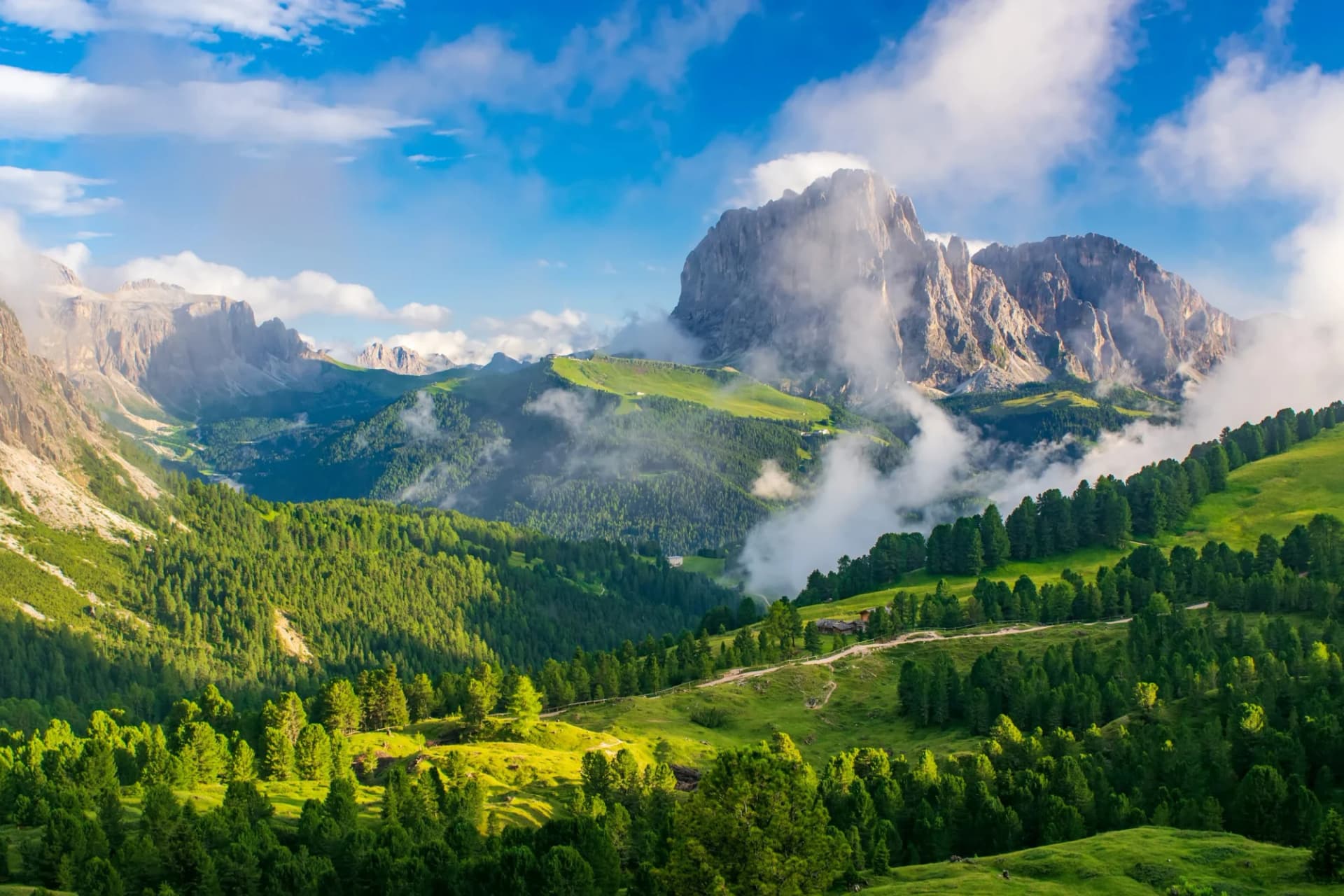 Sassolungo massif and Gardena Valley in Dolomite Alps, Italy, with green slopes and low clouds.