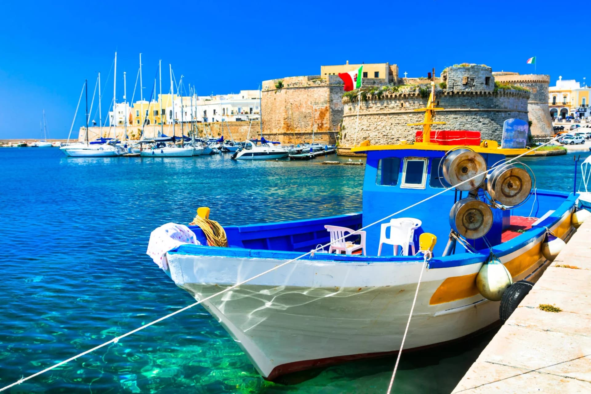 Blue and yellow fishing boat docked in harbor near historic castle in Puglia, Italy.