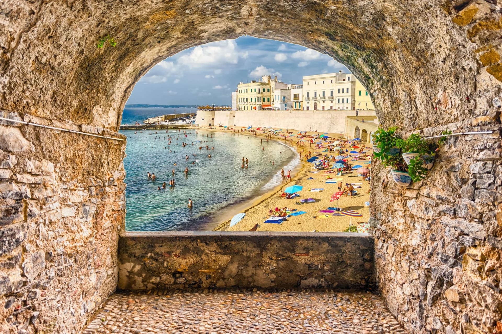 Beachgoers swimming and sunbathing viewed through a stone archway overlooking Gallipoli waterfront, Salento.