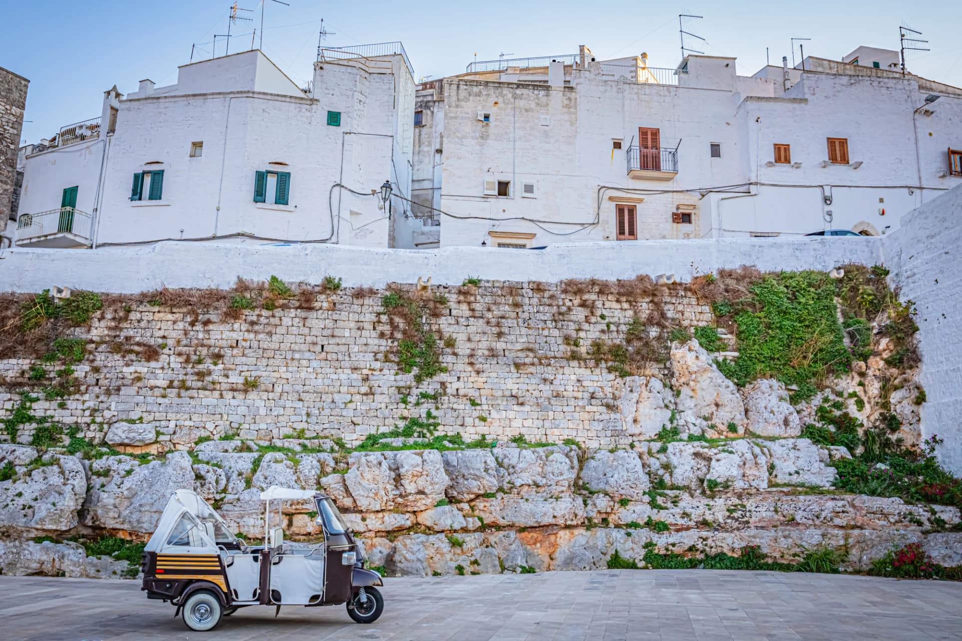 Tuk-tuk parked below white buildings and a stone retaining wall in Ostuni, the White City.