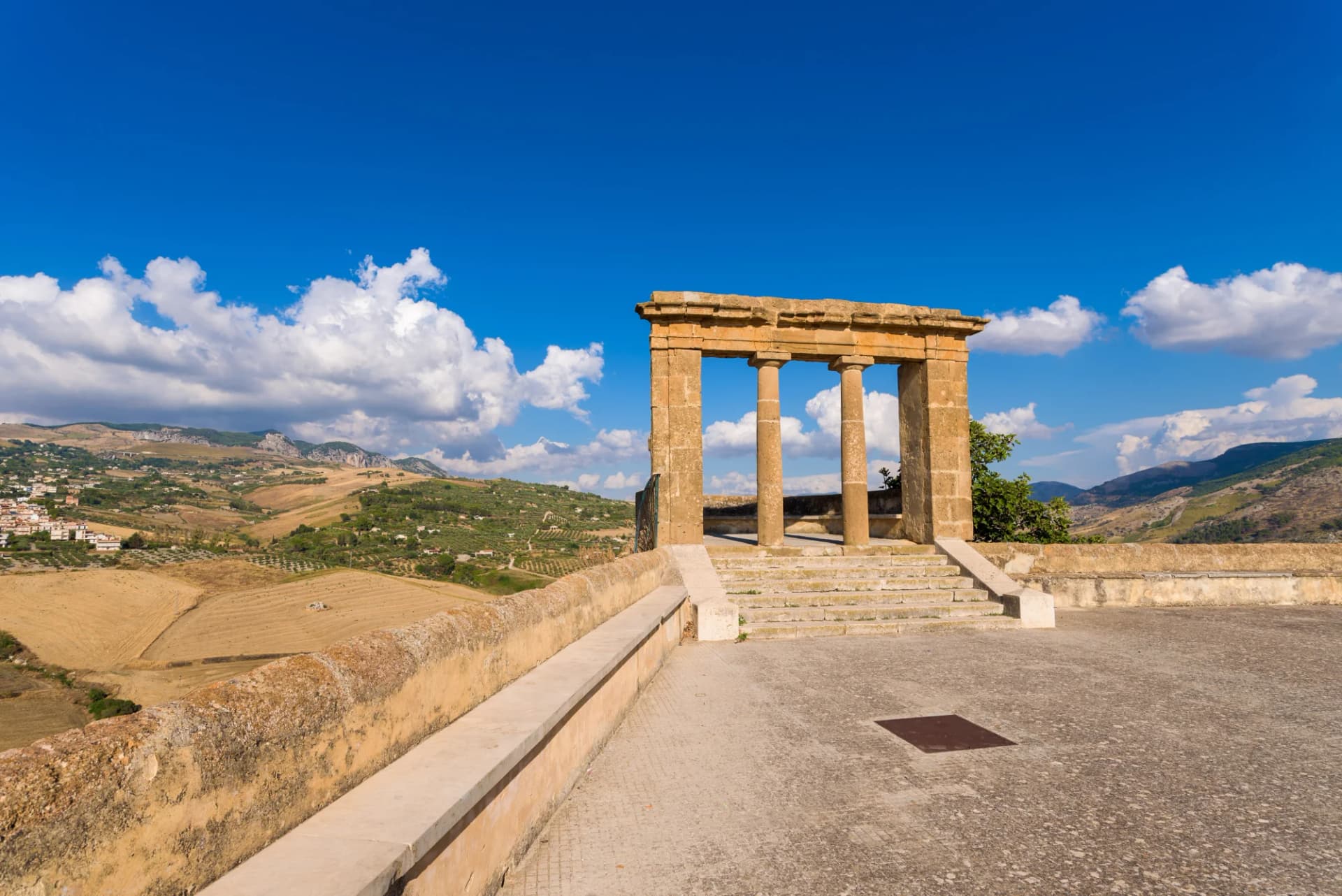 Terrazzo lookout with ancient columns overlooking a restored village and dry, rolling hills under a blue sky.
