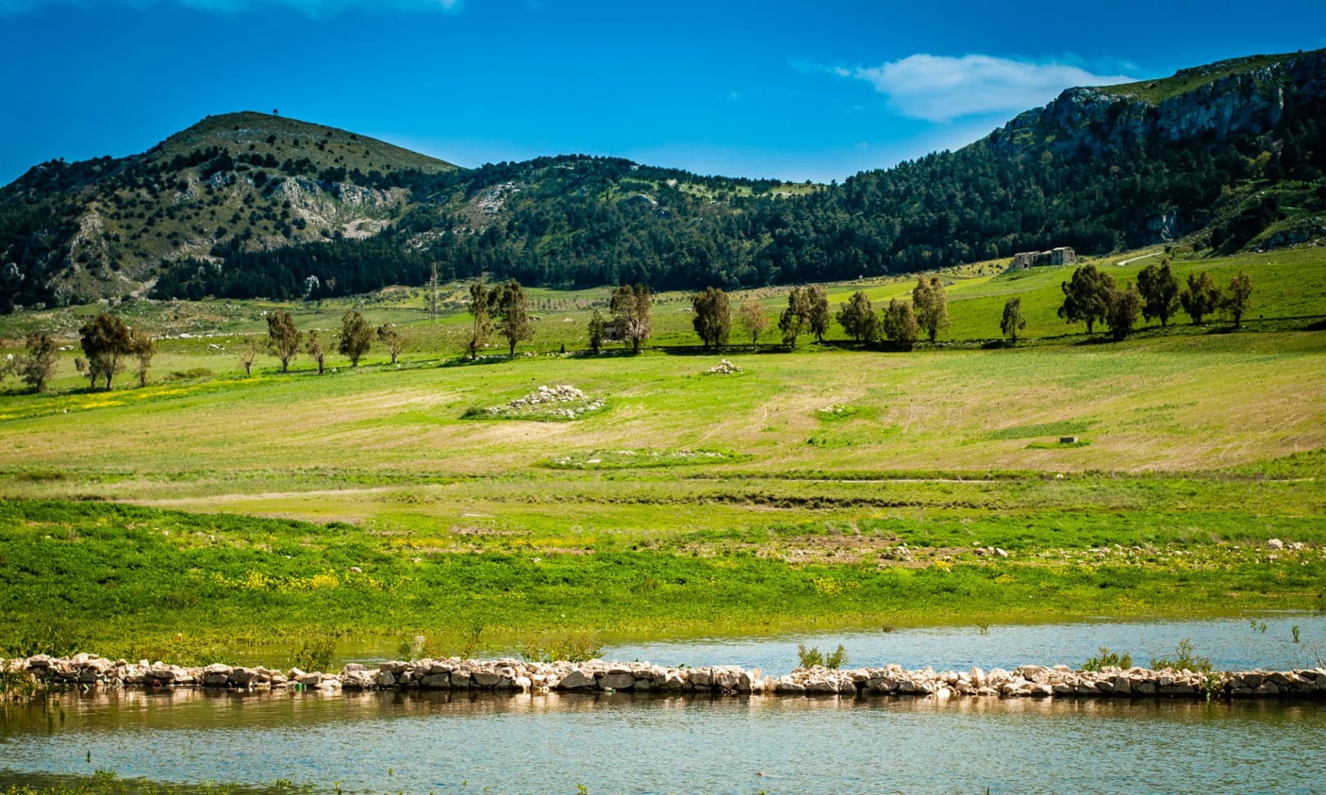 Lake Piana degli Albanesi reservoir with green hills and mountains under a blue sky in Palermo.