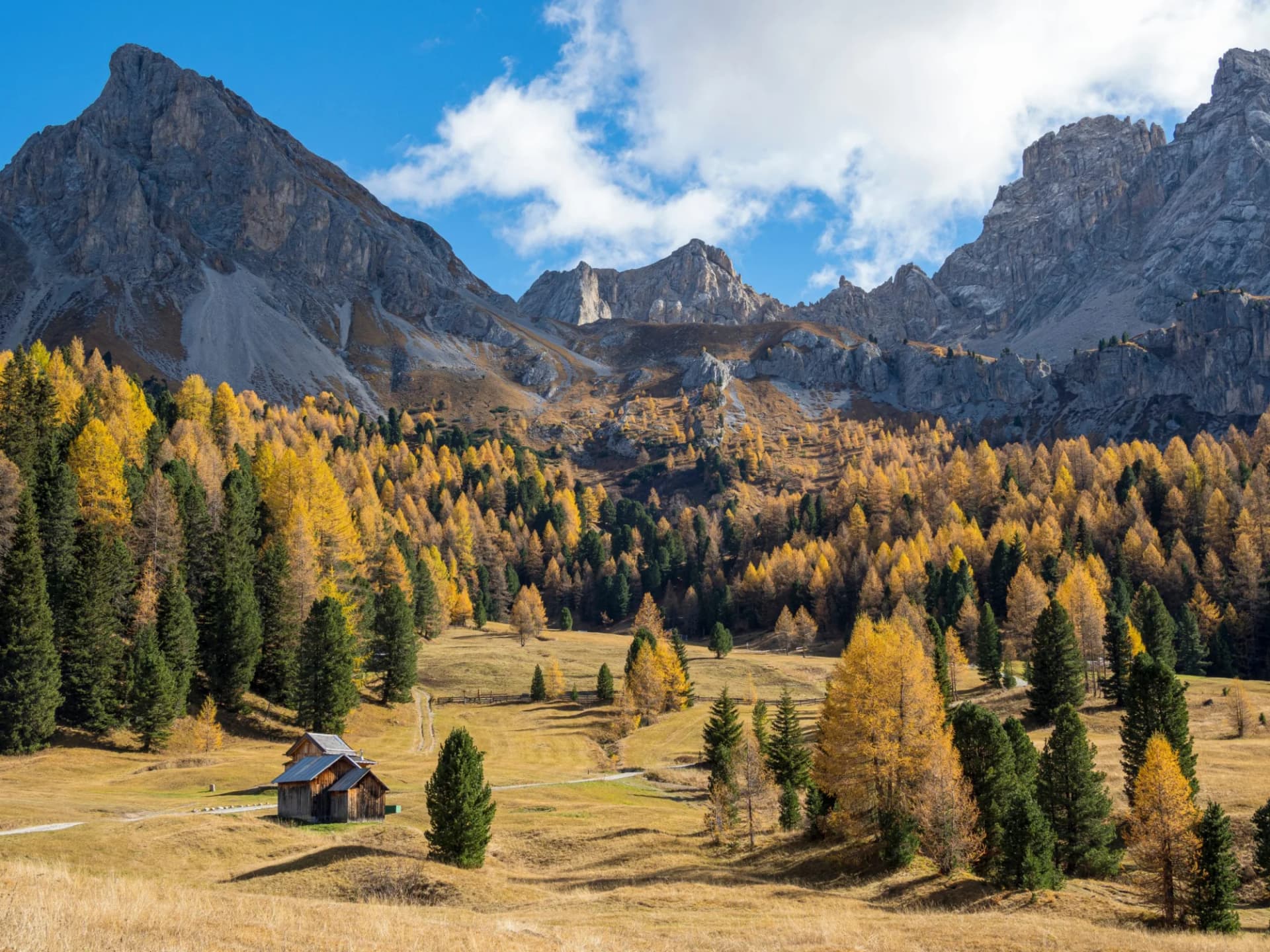 Alpine valley with wooden huts, yellow autumn trees, and rugged mountains under a blue sky.