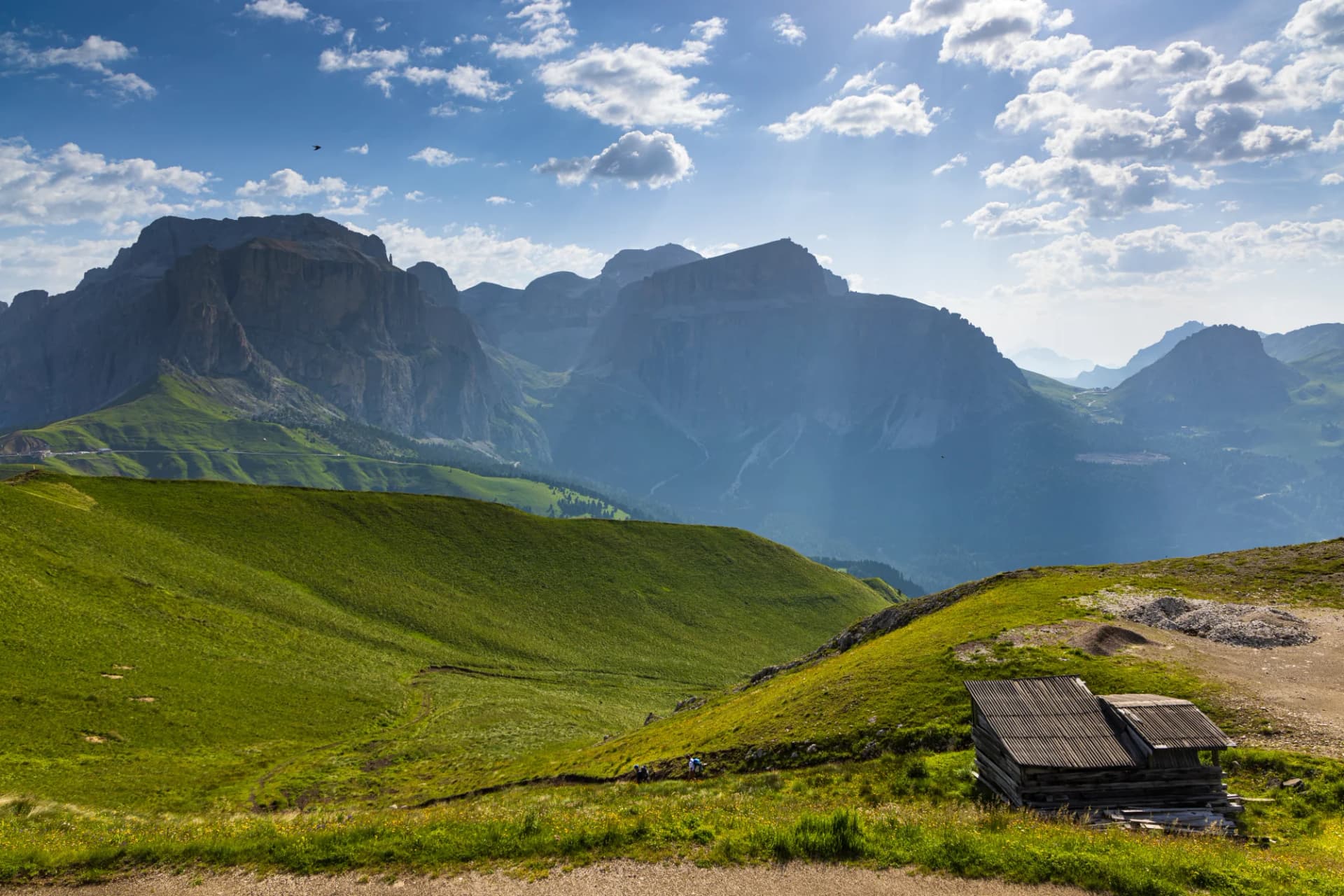 Alpine meadow with wooden hut, hikers, and dramatic mountain range under blue sky in Val di Fassa.